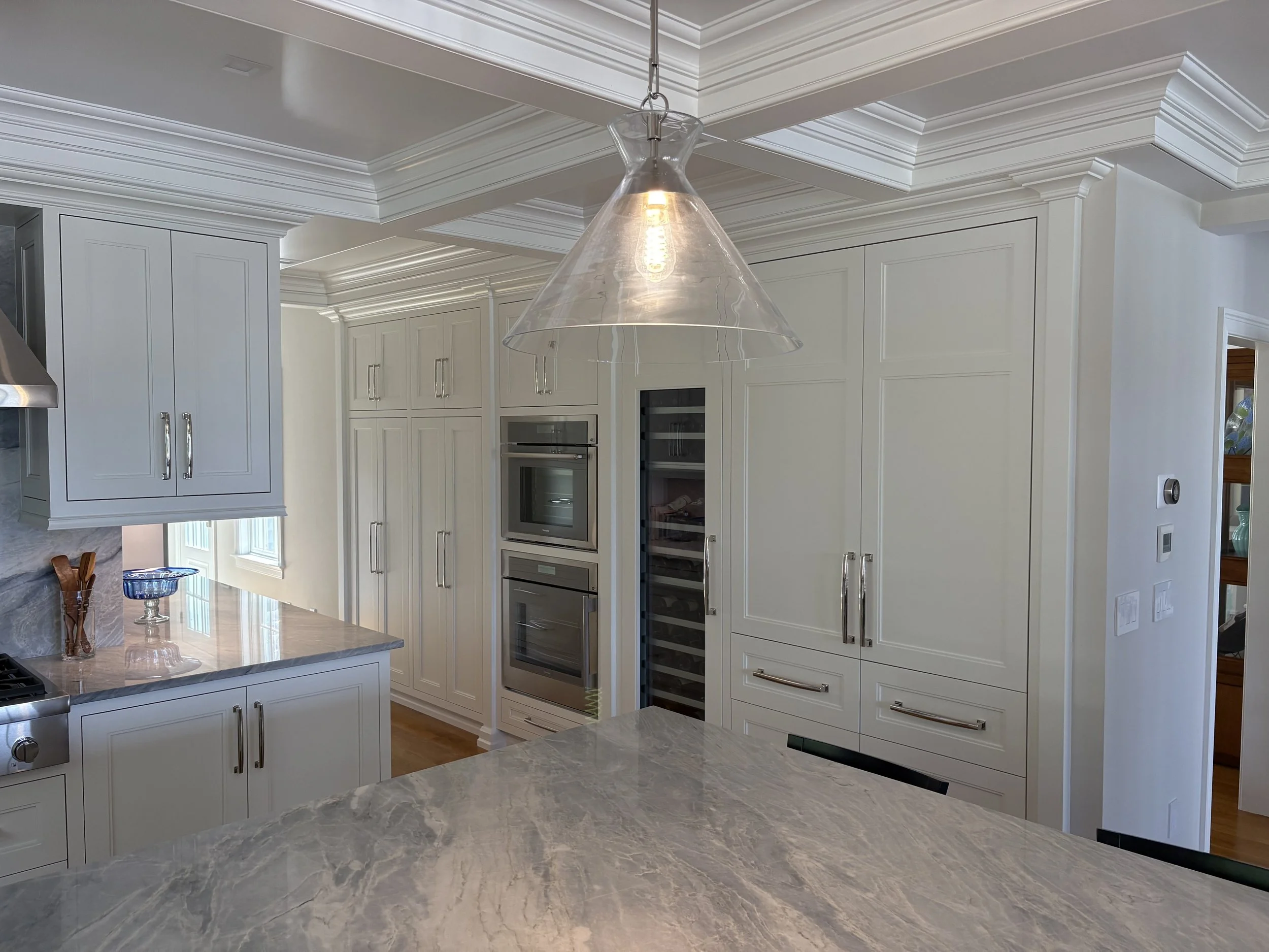Modern kitchen with white cabinetry and marble countertops, featuring built-in ovens and a glass pendant light