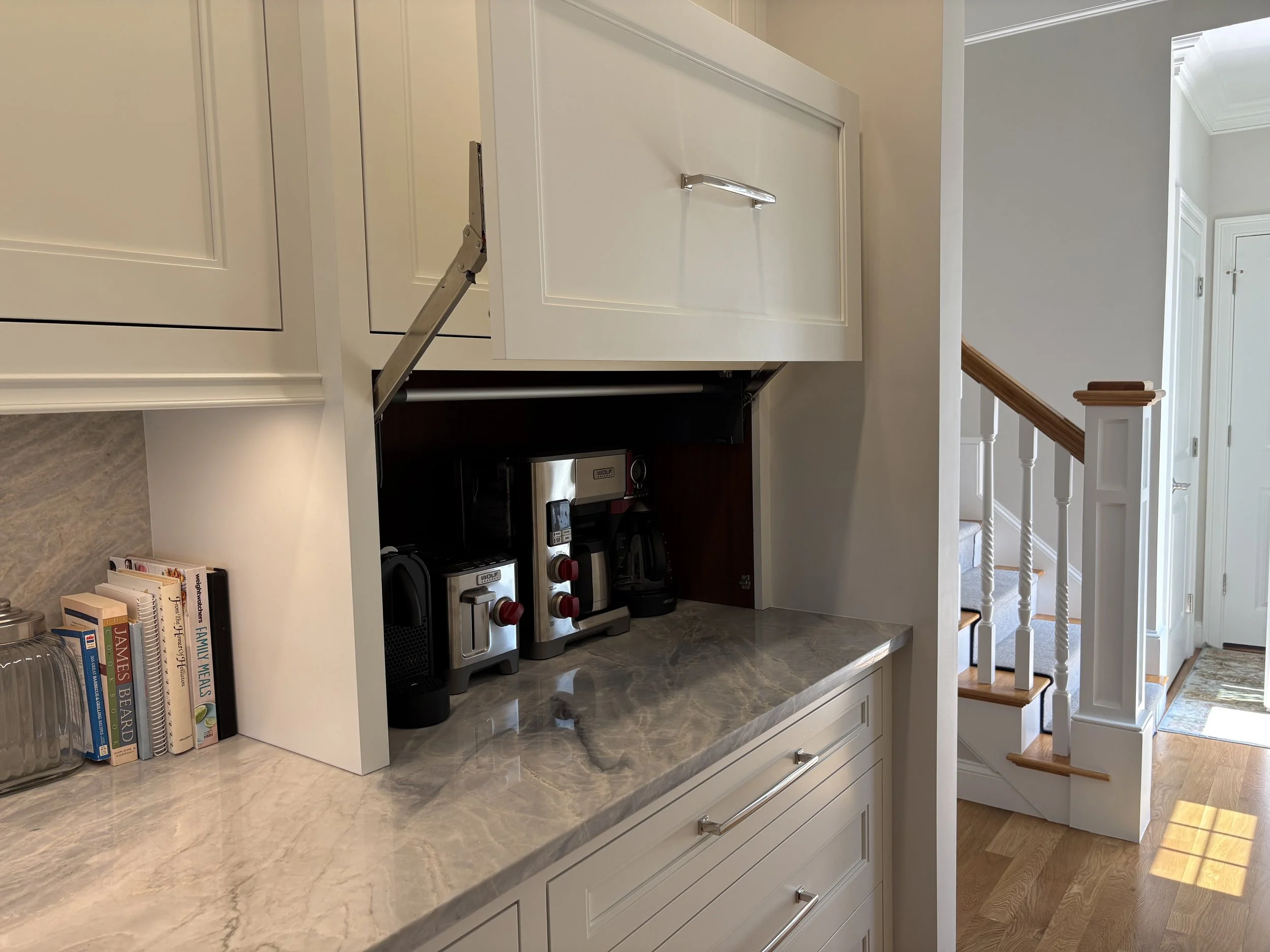 Kitchen counter with coffee makers, books, and decorative items, adjacent to staircase and hallway in a well-lit home.