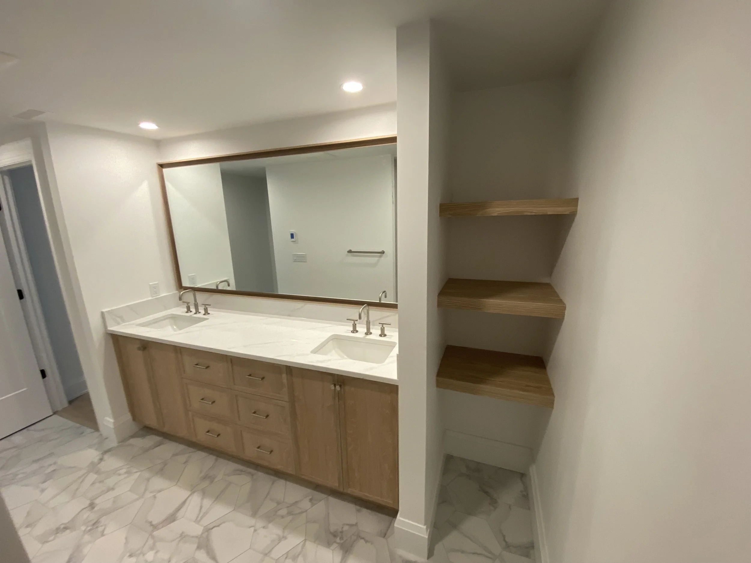 A bathroom vanity with two sinks, a large mirror, wooden cabinets, and three wooden shelves on the adjacent wall.