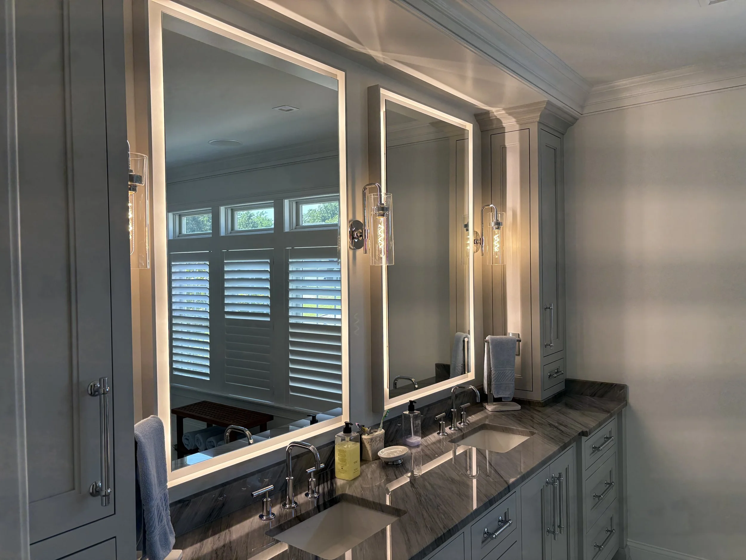 A modern bathroom vanity with two sinks, large illuminated mirrors, and clouds of toiletries. Two wall-mounted lights illuminate the area, and a window with white shutters is reflected in the mirrors.