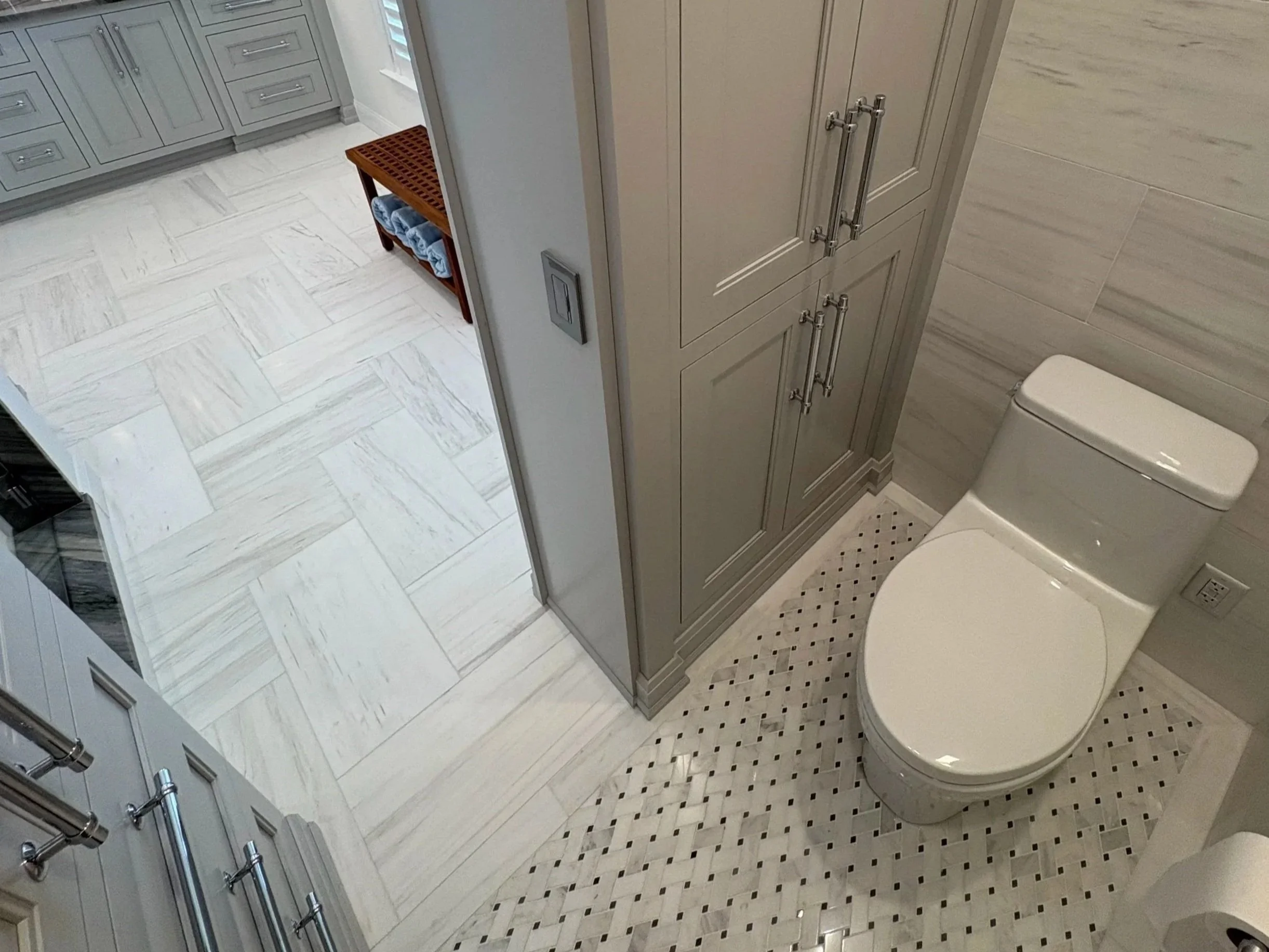 View of a bathroom with a toilet, gray cabinetry, and beige/white tile floors. There is a small wooden bench with blue towels on it.