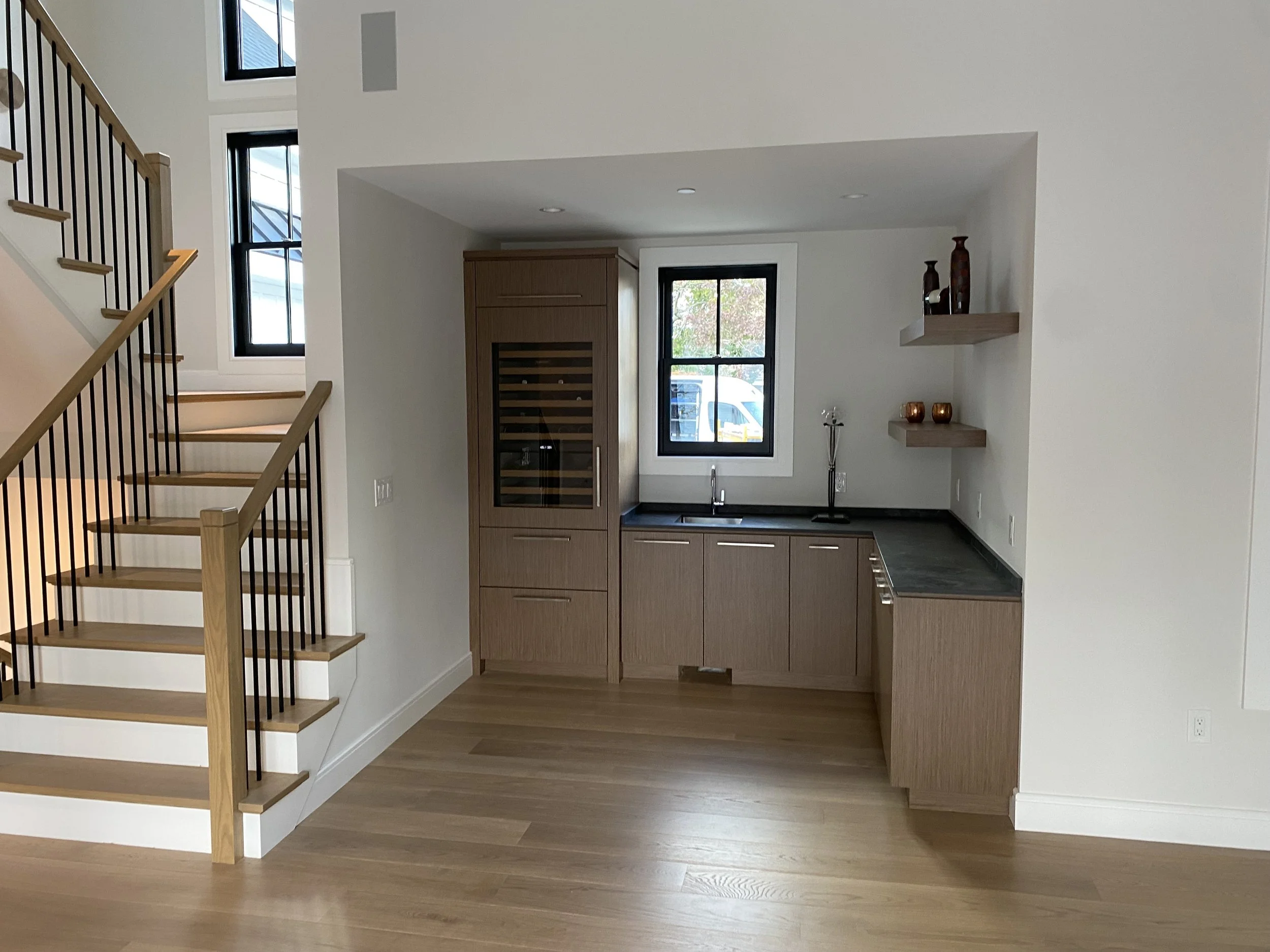 A modern kitchen with light wood cabinets, black countertops, a small sink, and floating shelves with decorative vases. There are two black-framed windows, one above the sink and one to the left, with trees and a parked vehicle outside. To the left i