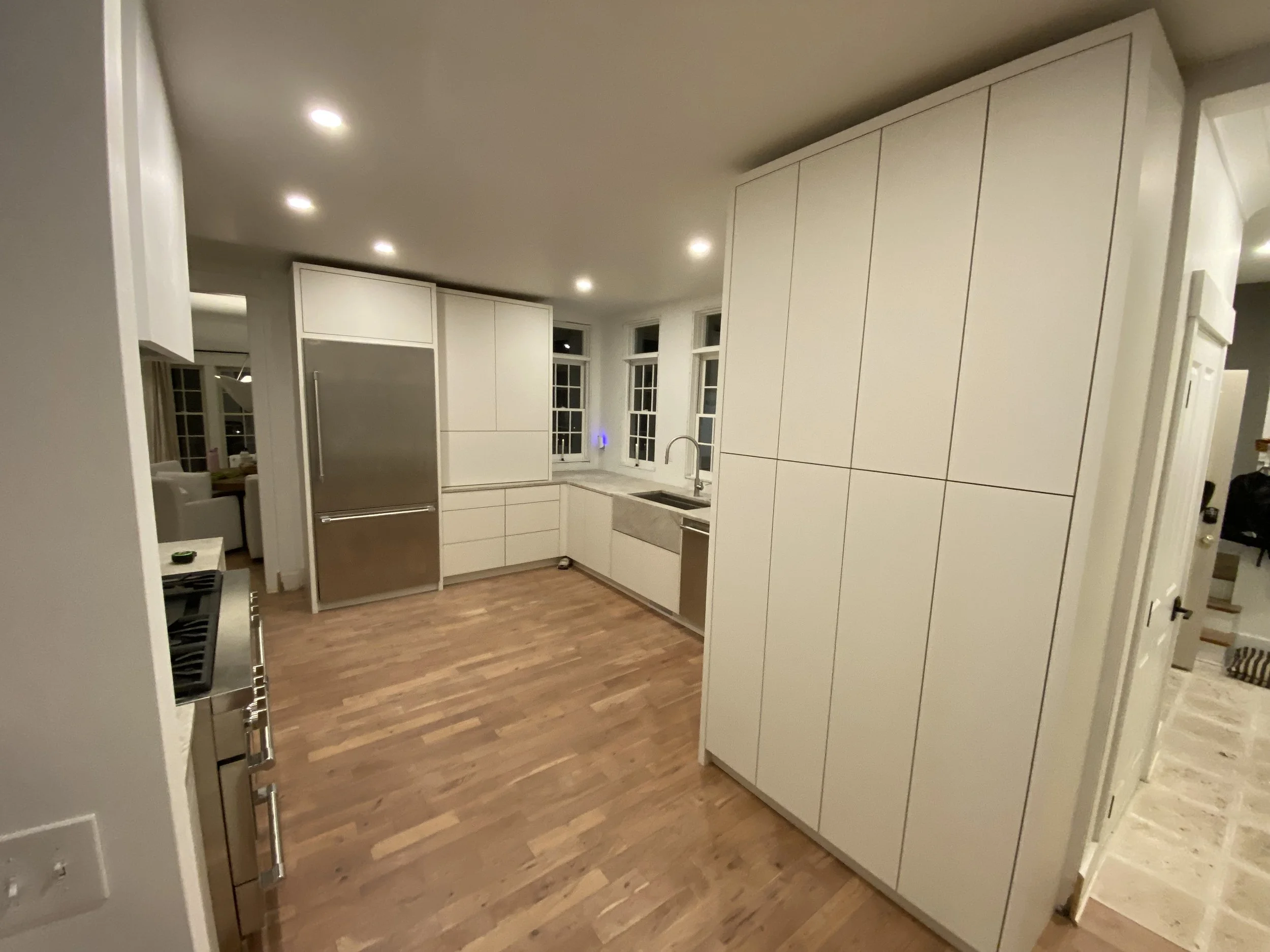 Modern kitchen with white cabinets, stainless steel refrigerator, hardwood floors, and a farmhouse sink under three windows.