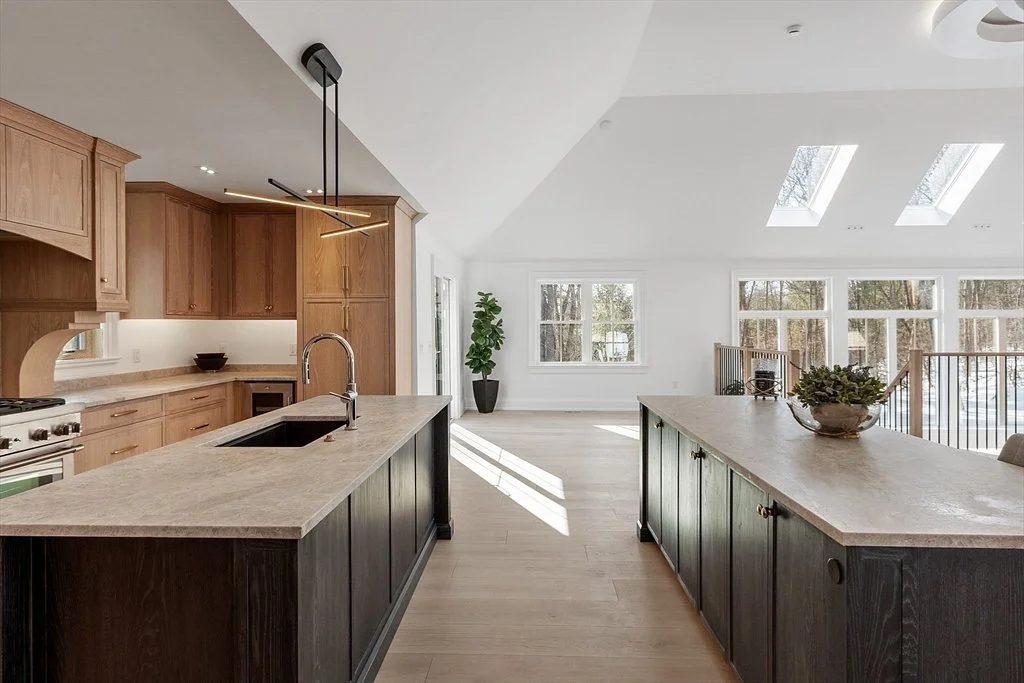 Open-concept kitchen and living area with wooden cabinets, light-colored countertops, and skylights. Large windows let in natural light, with a potted plant near the window and a decorative bowl with greenery on the central island.