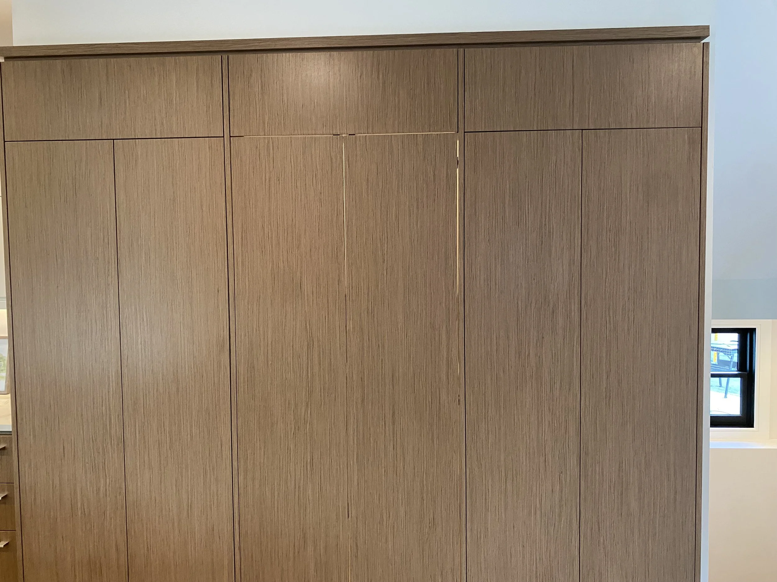 A large wooden closet or cabinet with multiple doors, situated next to a small window in a room with light gray walls.