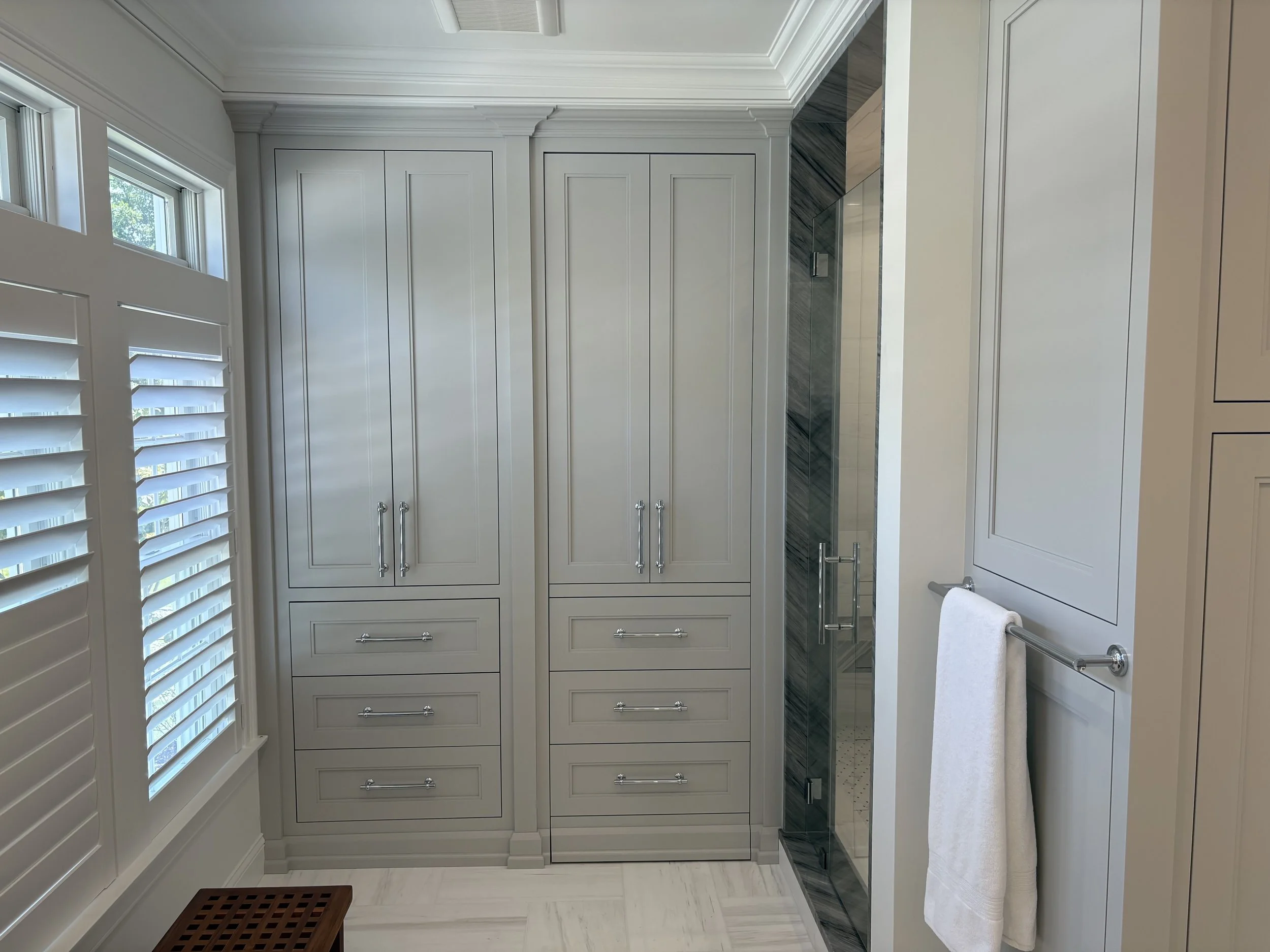 A modern bathroom with white cabinetry and a walk-in shower with a glass door. There are white plantation shutters on the windows and a towel hanging on a silver towel bar.