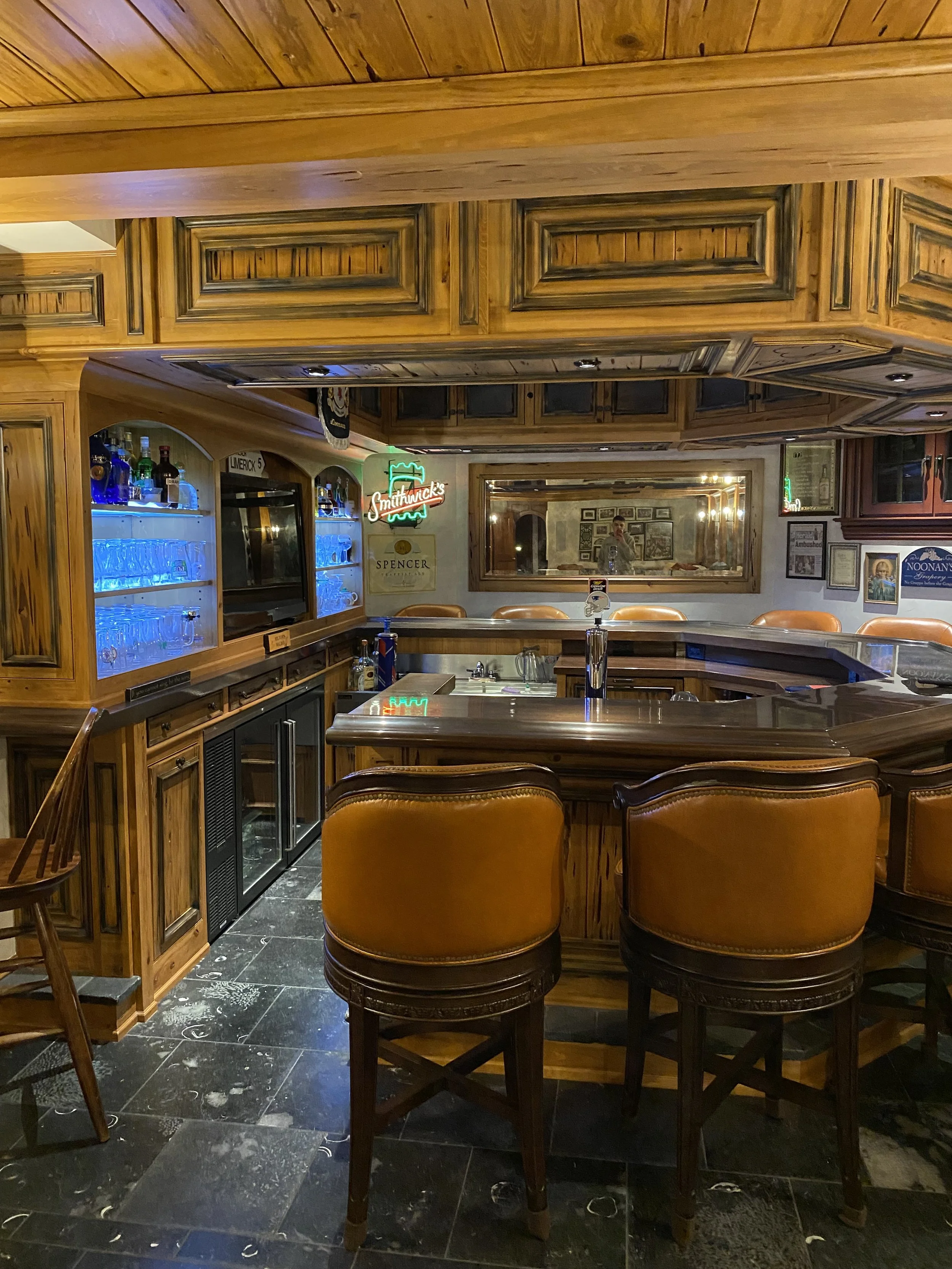 A cozy pub or bar with a wooden bar counter, leather chairs, and a mirror behind the bar. Various liquor bottles and glasses are on shelves, and neon signs decorate the wall.