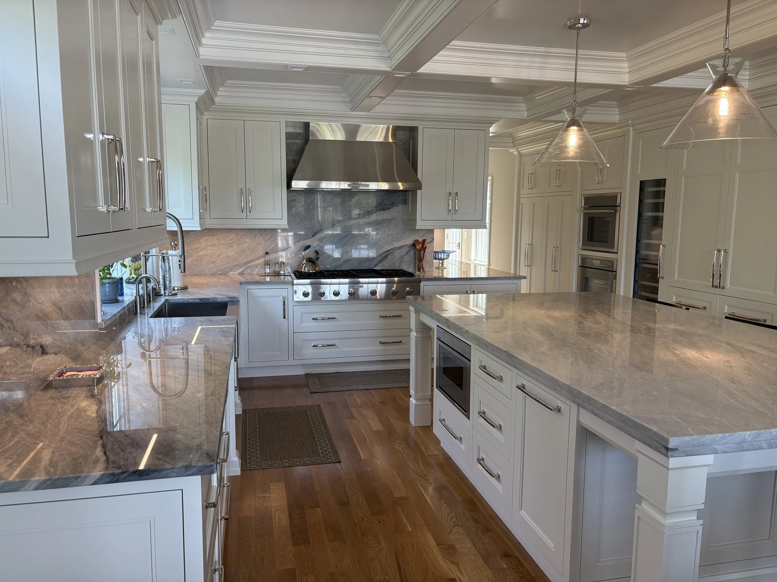 A spacious modern kitchen with white cabinets, a large marble island, hardwood floors, stainless steel appliances, pendant lighting, and a marble backsplash.