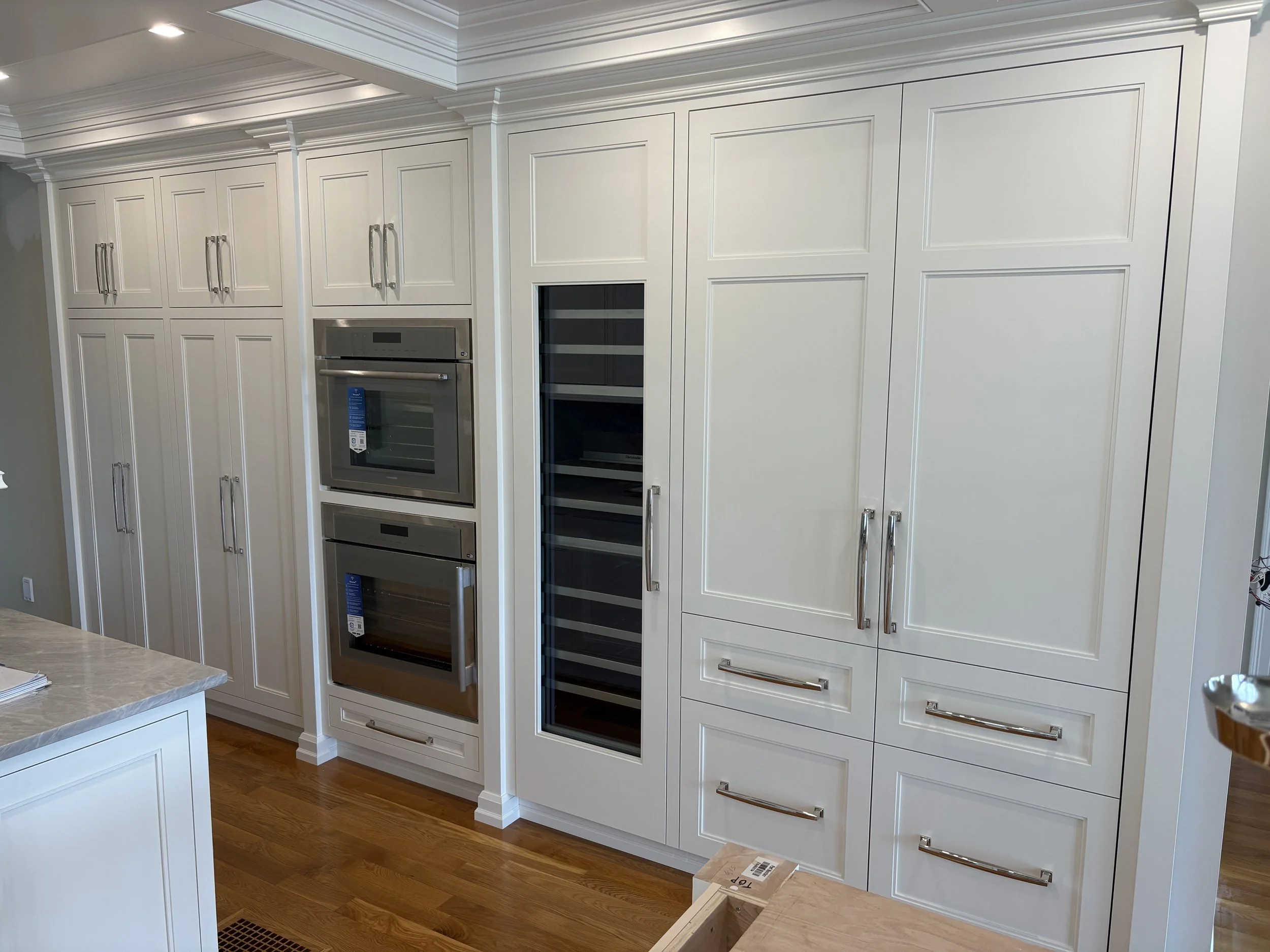 White kitchen cabinets with built-in double oven, wine cooler, and multiple drawers, hardwood floor, marble countertop in foreground.