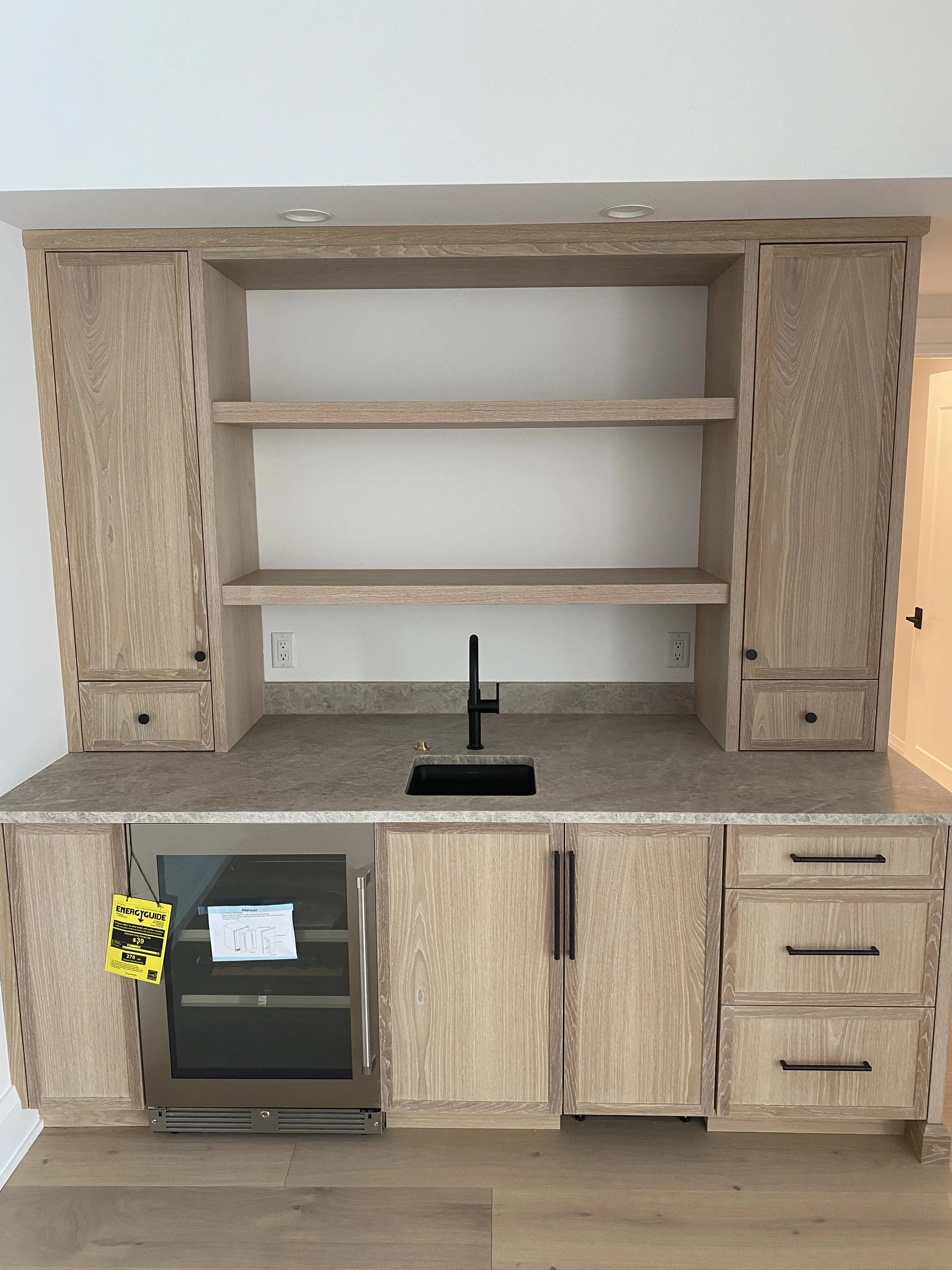 Kitchen area with light wood cabinets, a small built-in oven with an energy guide tag, a black faucet at a small sink, and open shelving above the counter.