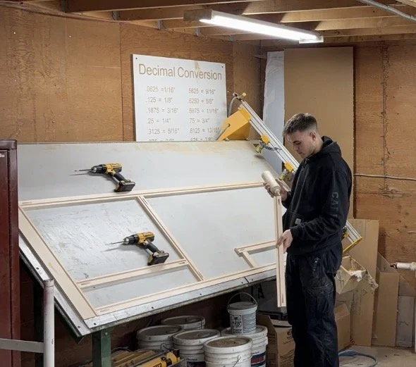 A young man in a black hoodie and pants working on a woodworking project in a workshop with wooden walls and ceiling. He is holding a piece of wood and standing next to a large angled workbench with clamps. There are buckets underneath the workbench and a projector screen displaying a 