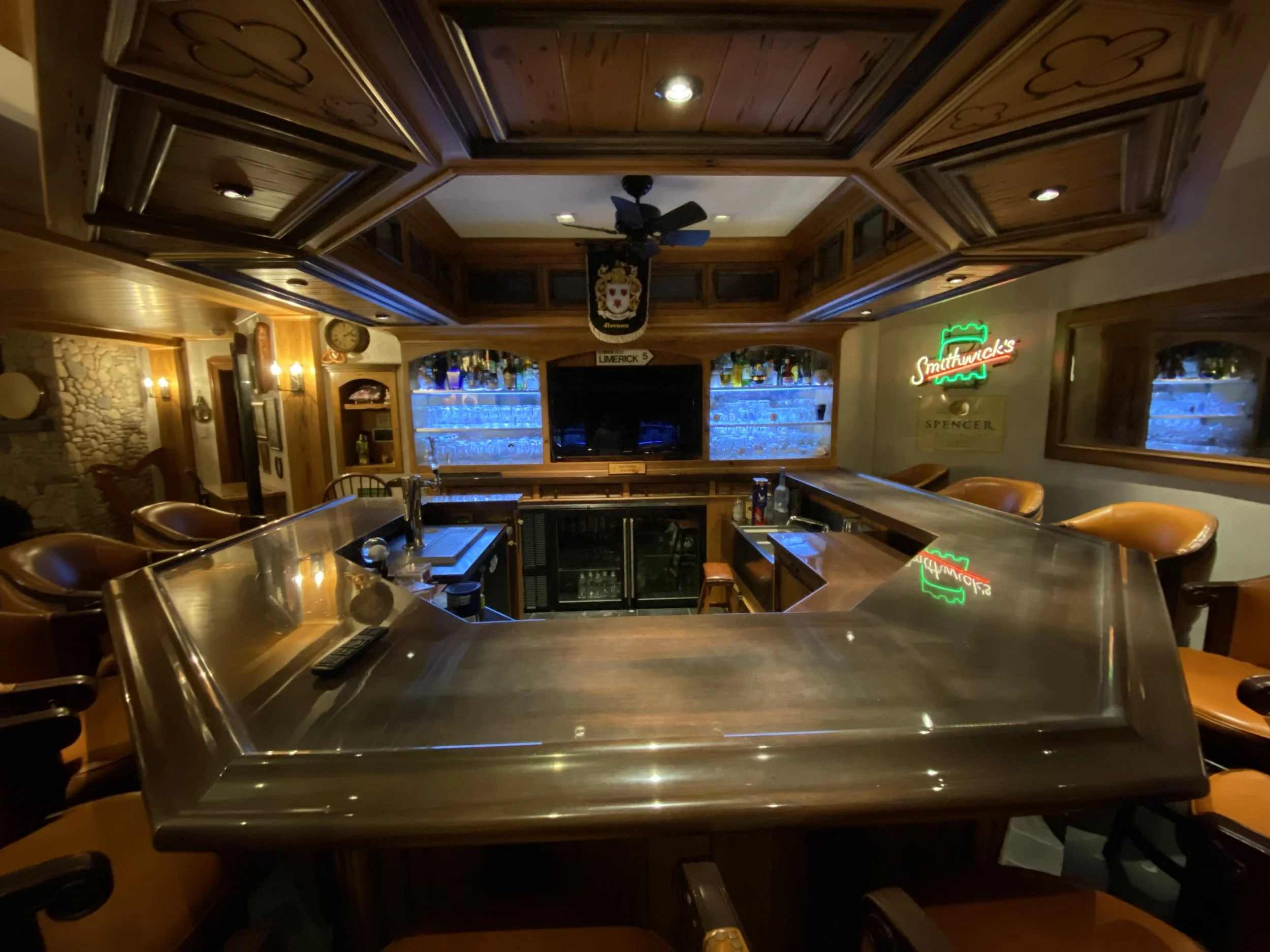 Empty bar with wooden decor, illuminated shelves with bottles, neon signs, and brown leather chairs around a curved bar counter.