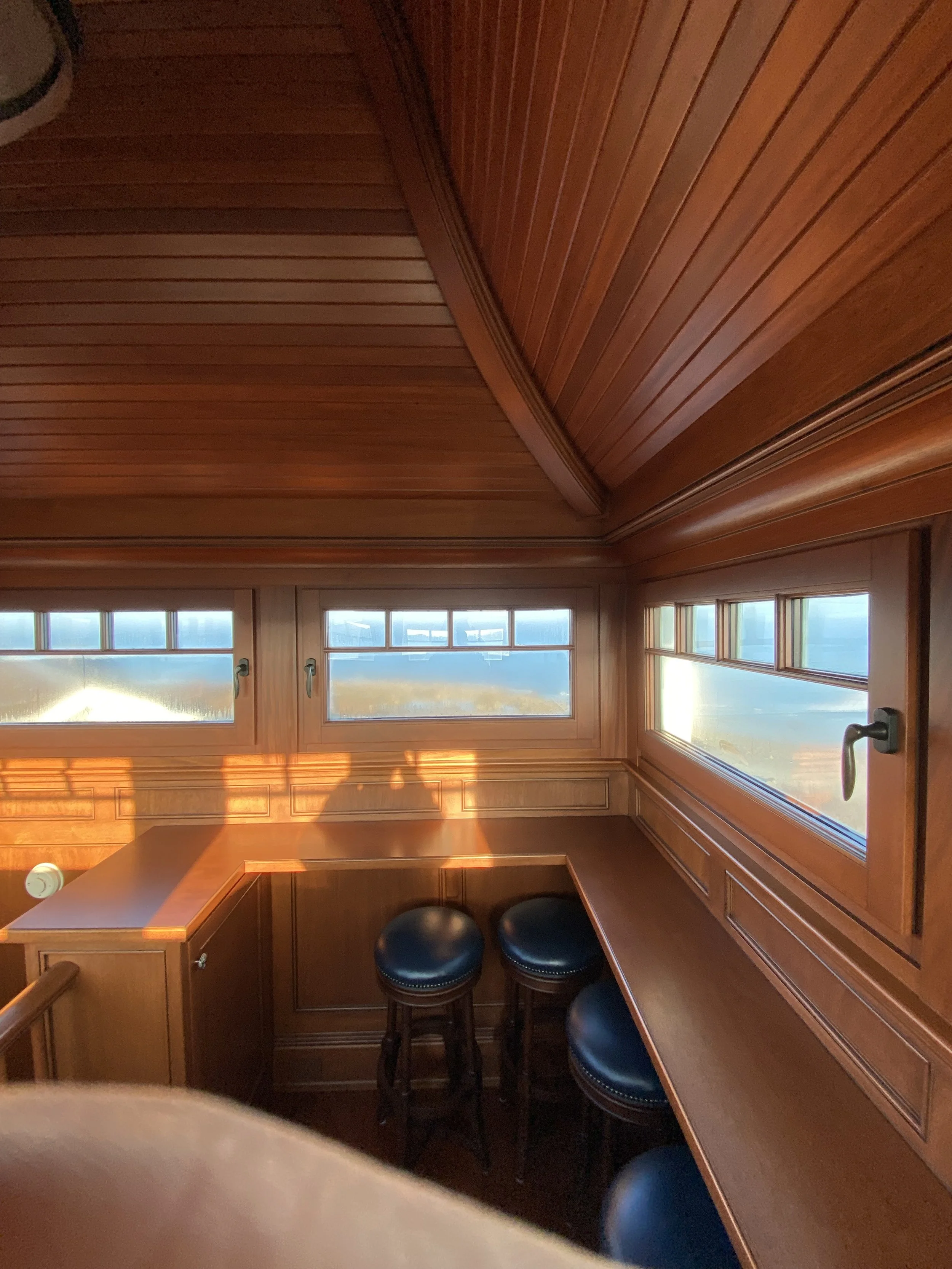 Sunlit cozy reading nook with wooden walls, ceiling, and built-in window seat with three black cushioned stools.