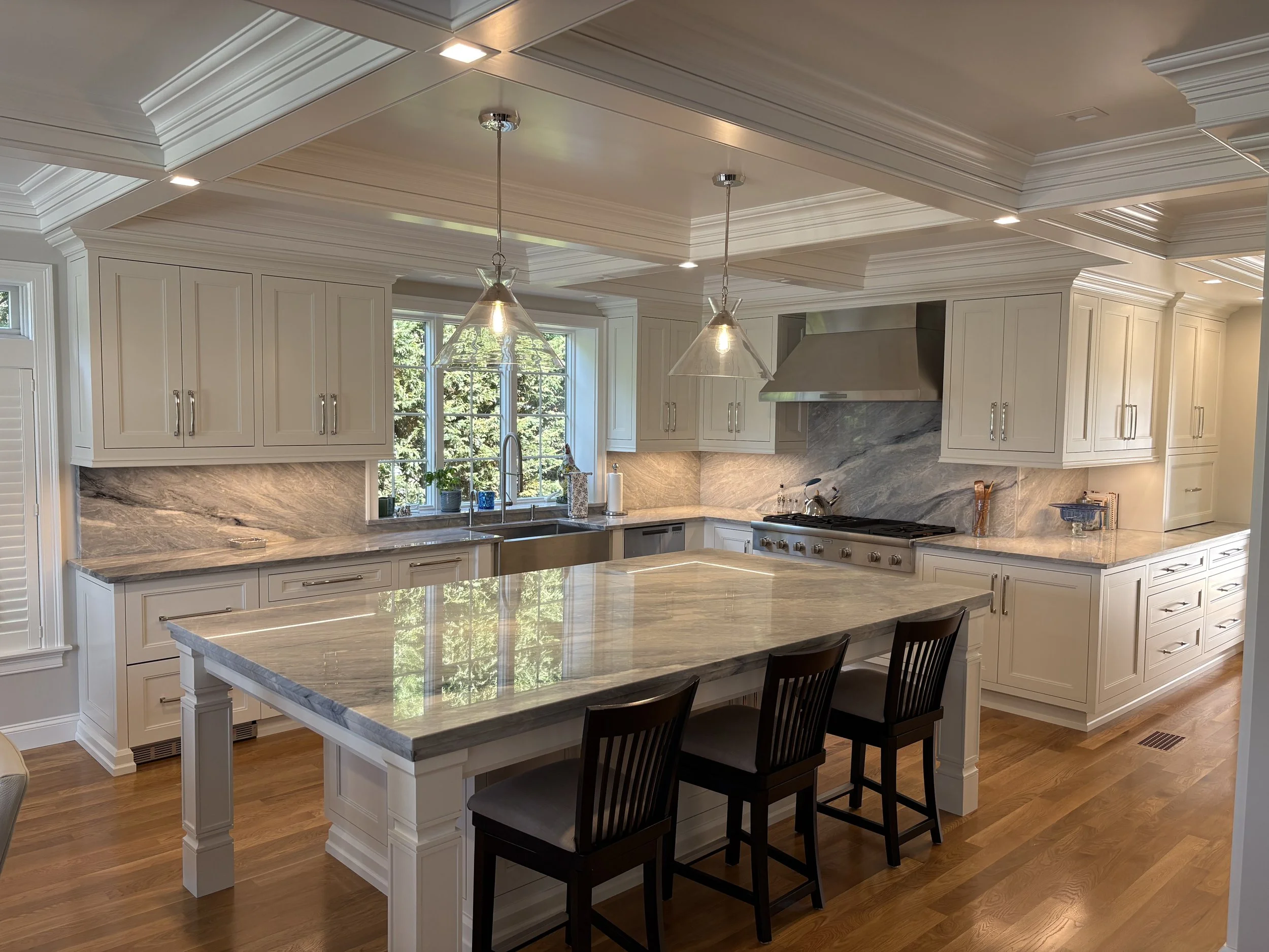 Modern kitchen with white cabinets, marble countertops, pendant lights, stainless steel range hood, and a large island with three chairs.