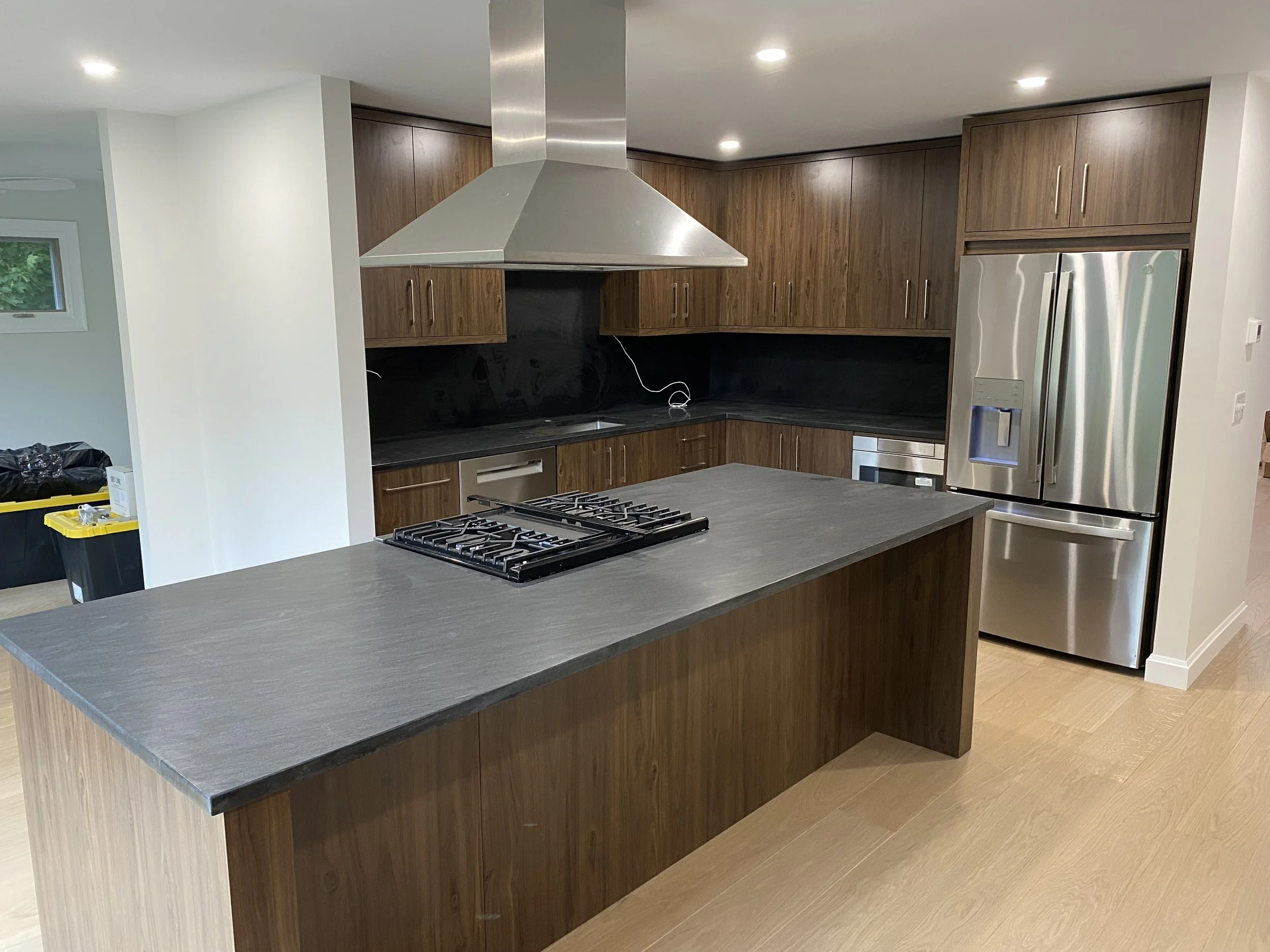 Modern kitchen with wooden cabinets, a stainless steel refrigerator, a gas stove on the kitchen island, and a range hood. There is a black backsplash and light wooden flooring.