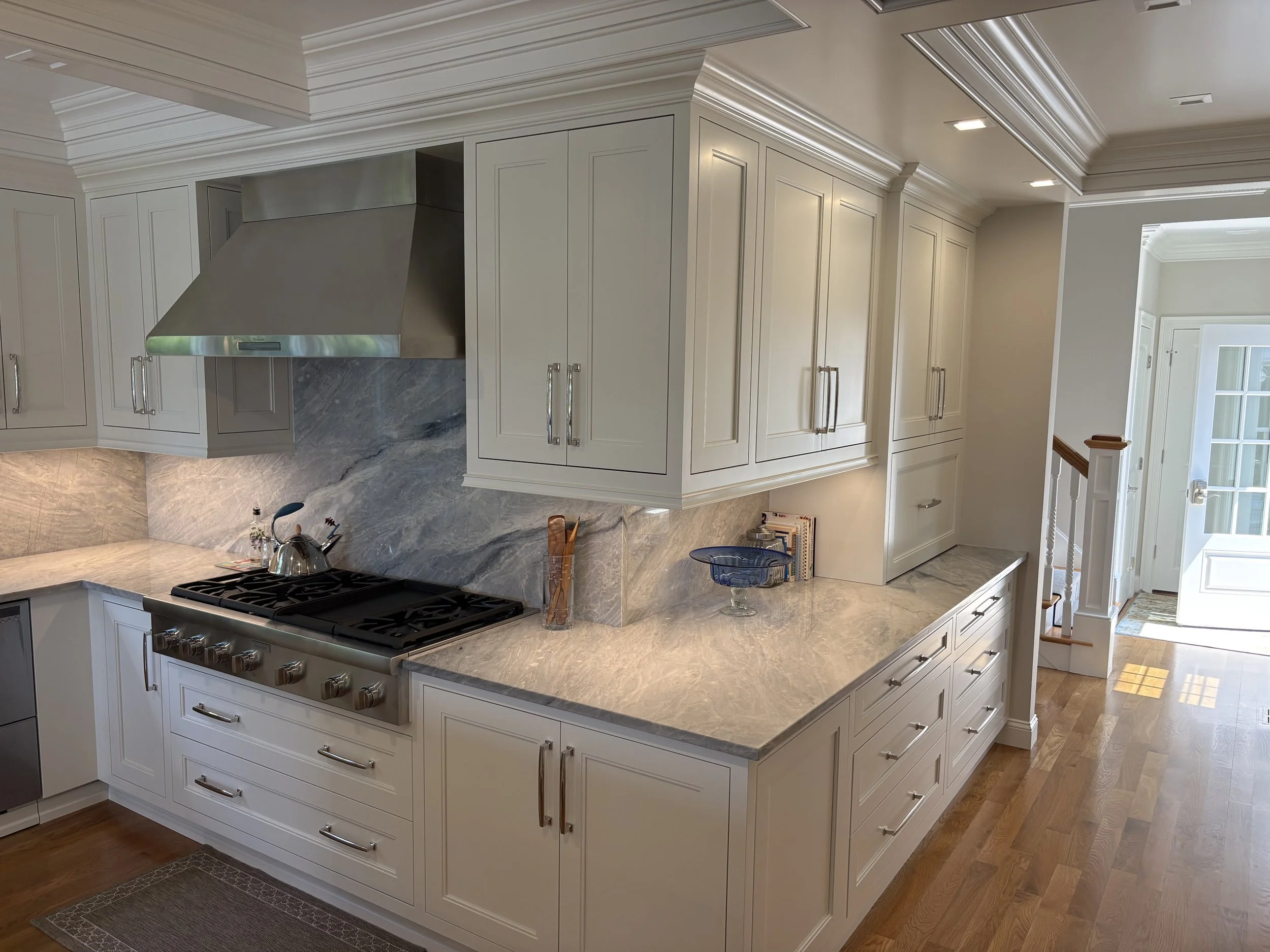 Modern kitchen with white cabinetry, marble countertops, stainless steel appliances, hardwood floors, and a view of a hallway with a staircase.