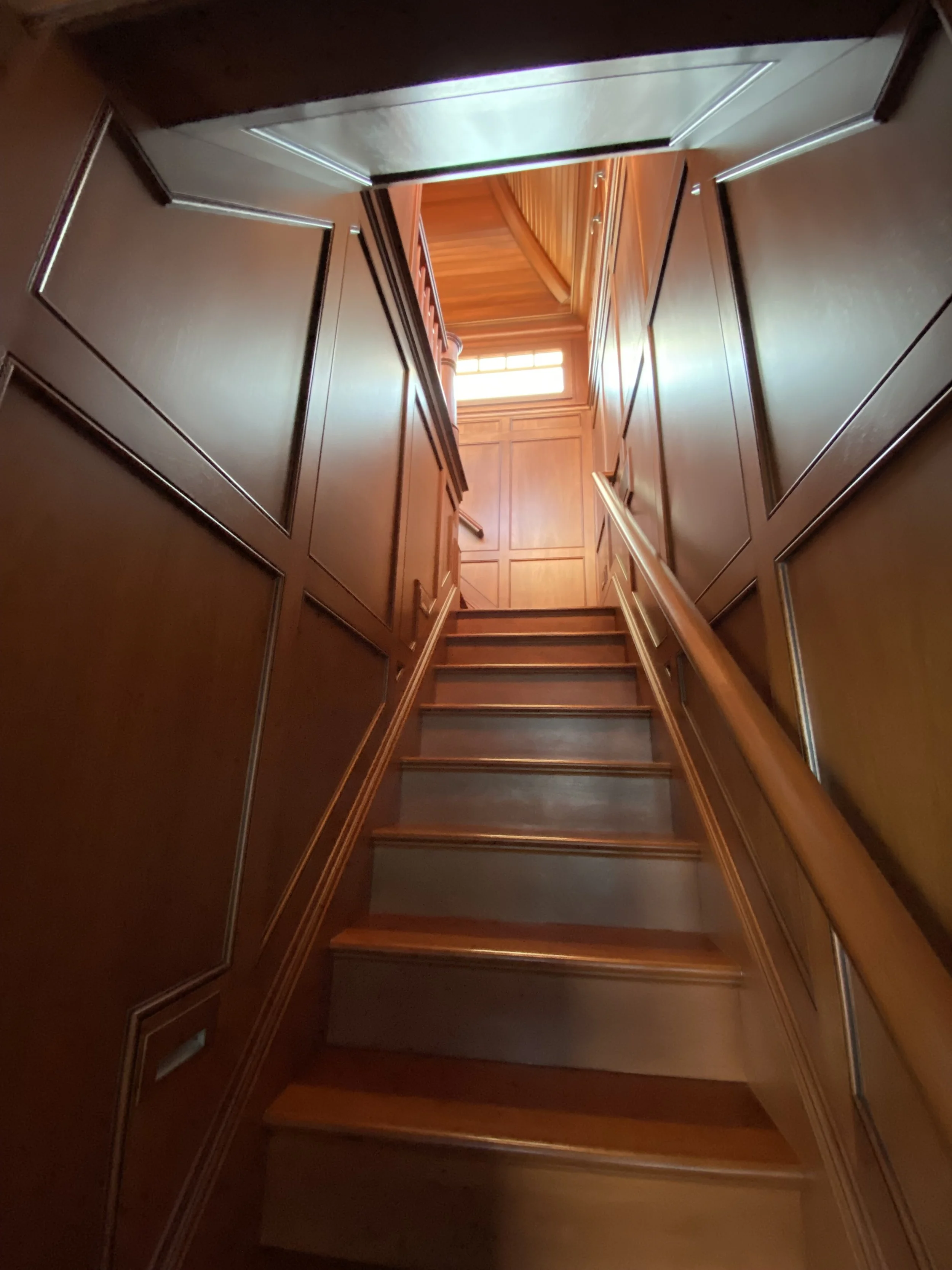 Wooden staircase leading up to a bright window with a wooden interior, viewed from the bottom of the stairs.