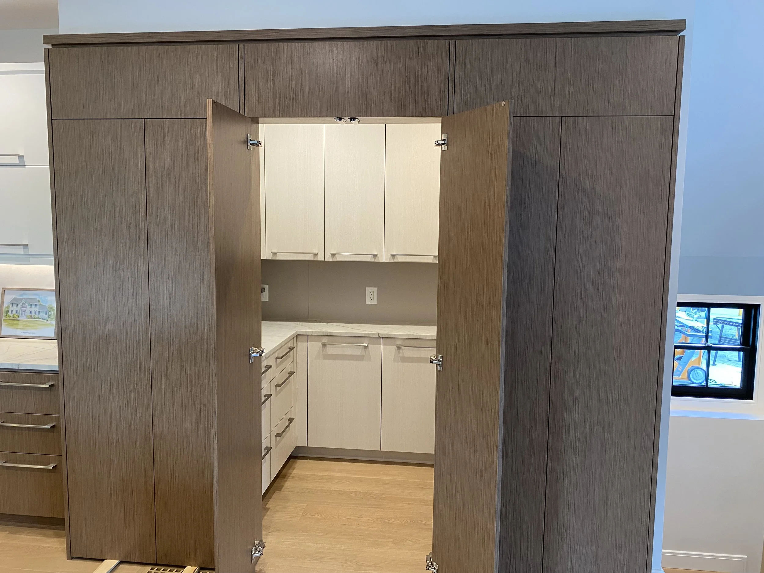 Open wooden cabinet doors revealing a small kitchen area with beige cabinets and countertop, against a gray wall with electrical outlets, and a window showing construction equipment outside.