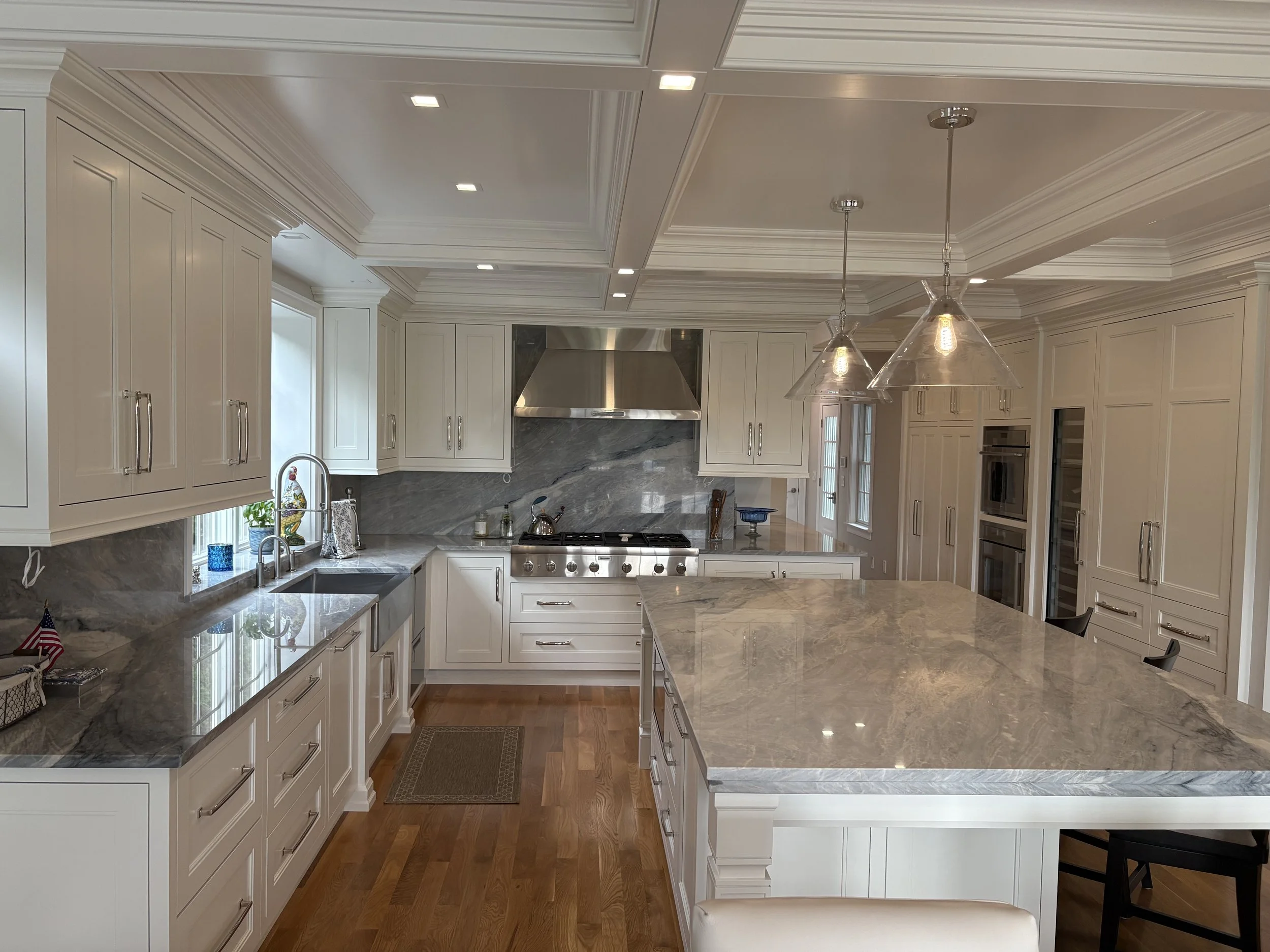 A spacious, modern kitchen with white cabinetry, marble countertops, hardwood floors, and stainless steel appliances. There are pendant lights hanging over the island and a window above the sink.