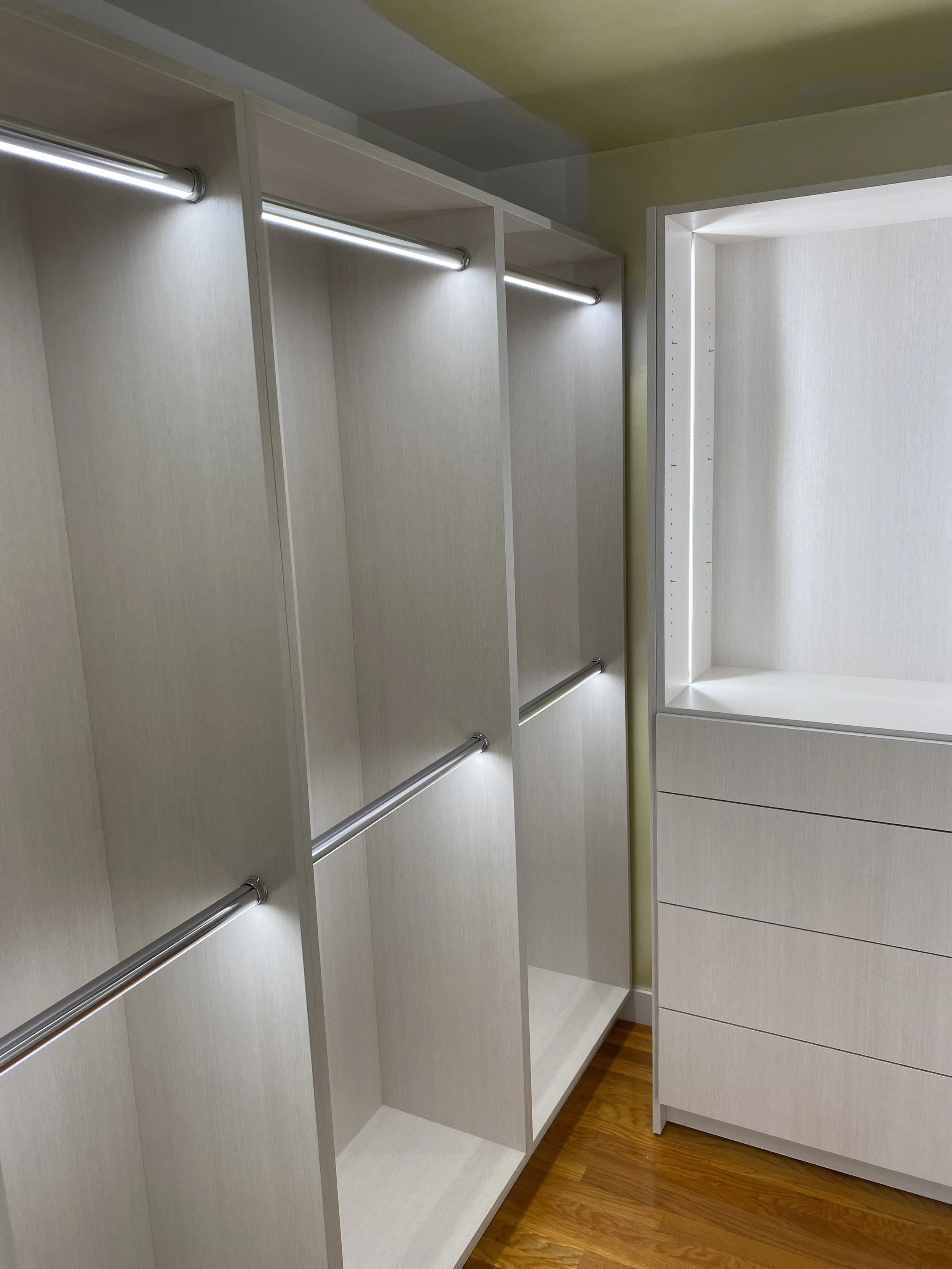 Empty white closet with hanging rods and built-in drawers, adjacent to a small storage cubby with shelves, on a hardwood floor.