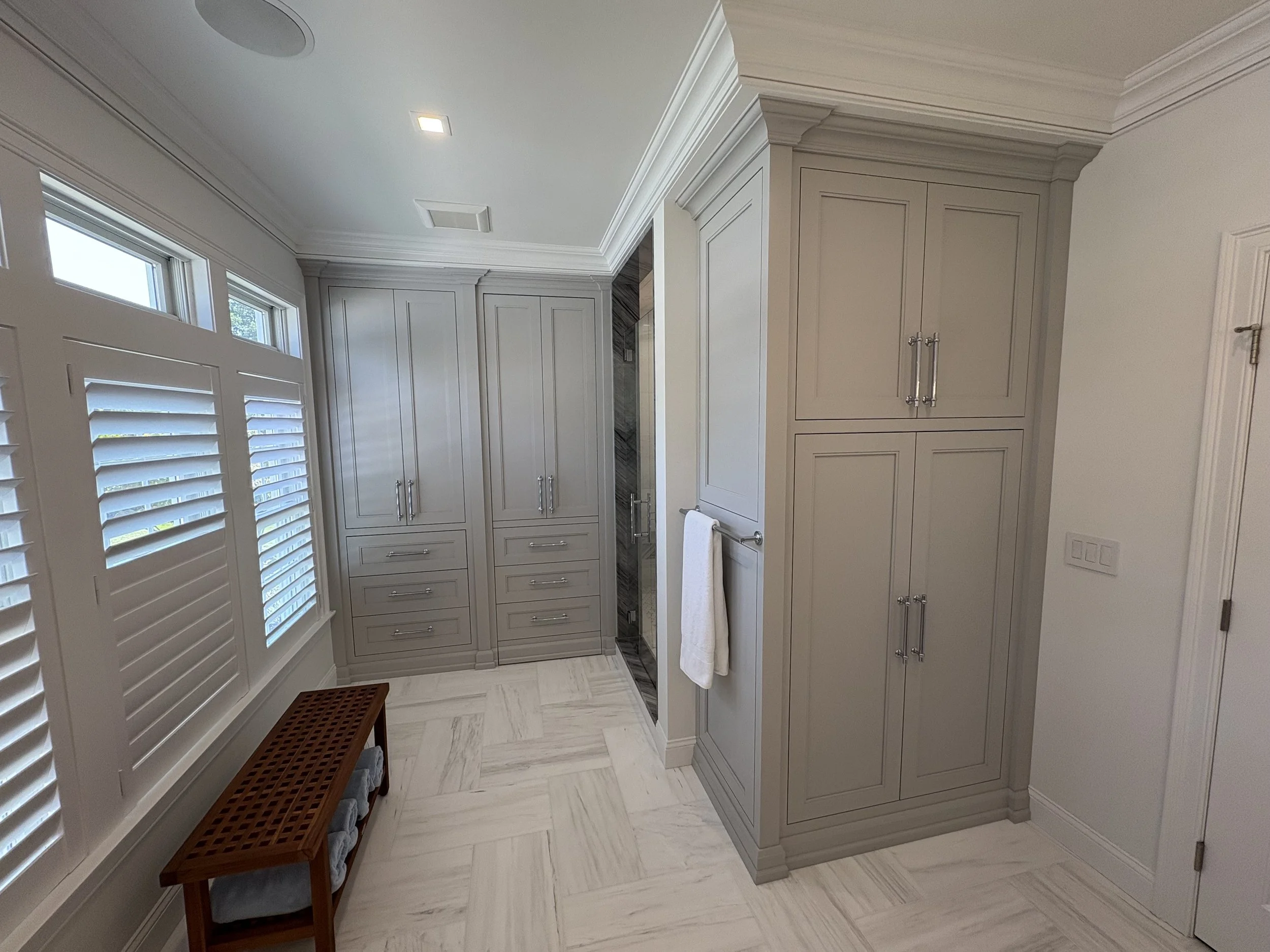 A bathroom corner with built-in gray cabinets, white crown molding, a window with white plantation shutters, a wooden bench with towels underneath, and a white towel hanging on a towel bar.