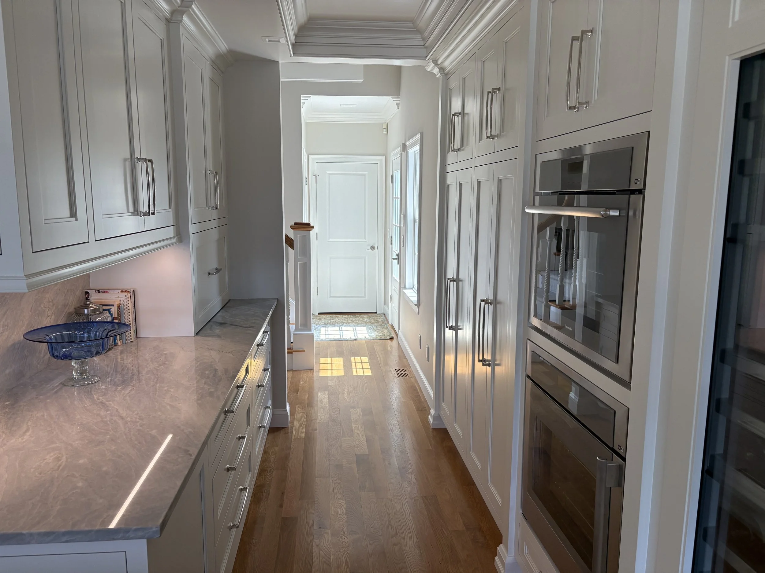 A narrow kitchen with white cabinets, a marble countertop, silver appliances, hardwood floors, and natural light from windows near the front door.