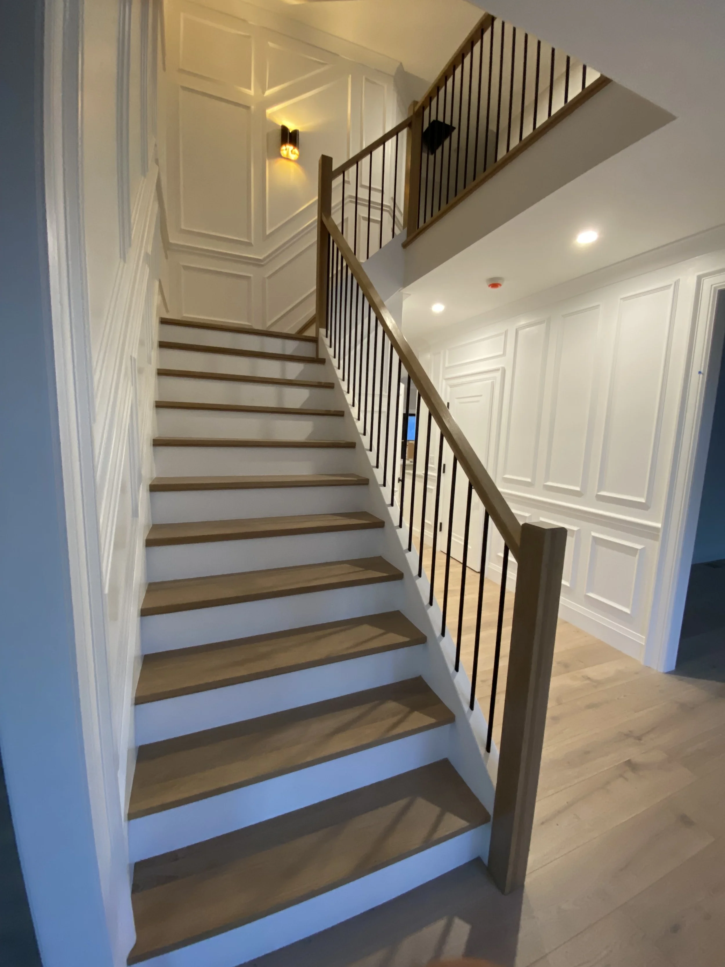 Interior staircase with wooden steps, white risers, black metal balusters, and a wooden handrail, leading to an upper floor with wall paneling and decorative wall sconces.