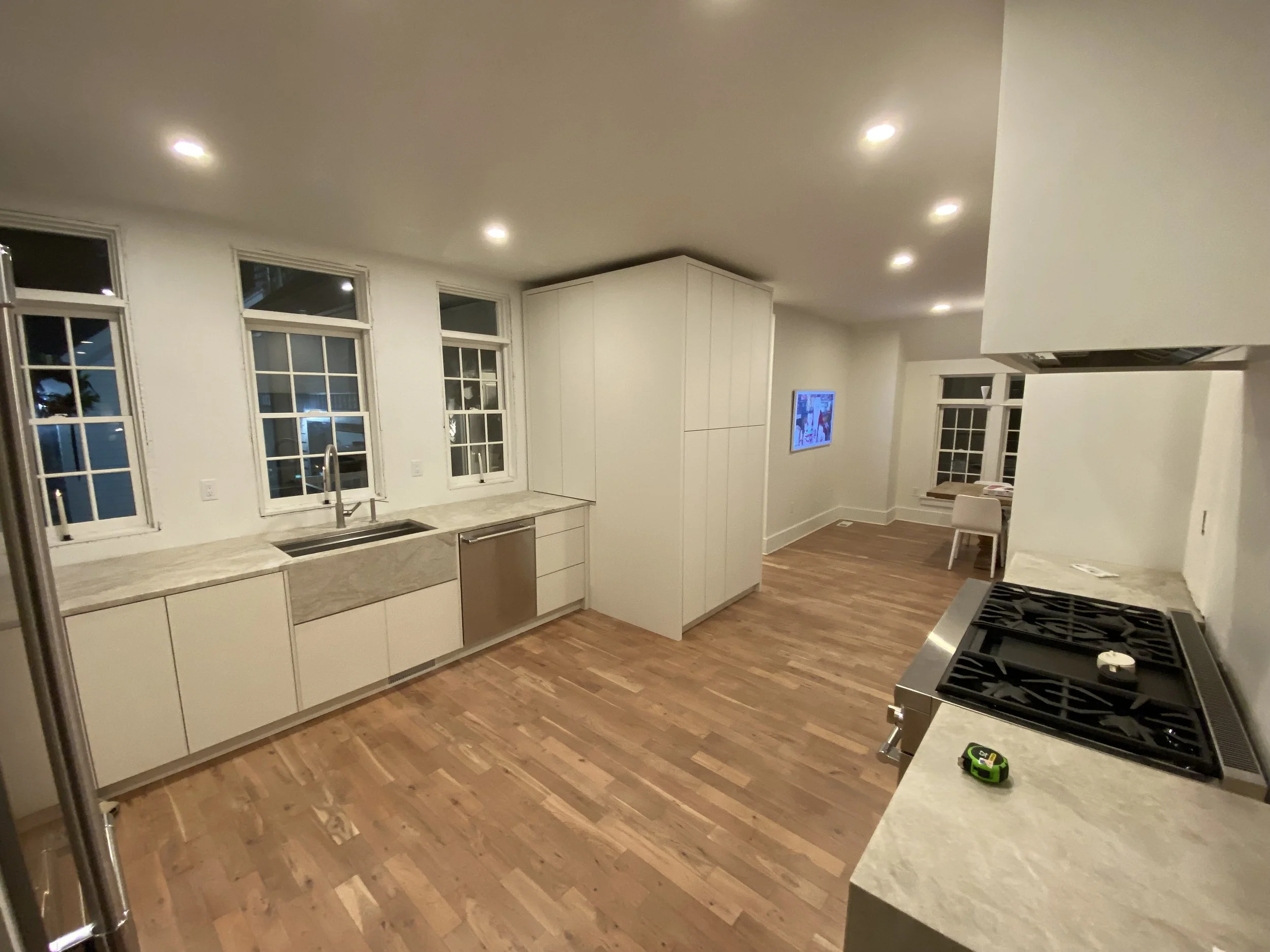 Modern kitchen with white cabinets, marble countertops, a stainless steel dishwasher, a gas stove, hardwood floors, and large windows at night.