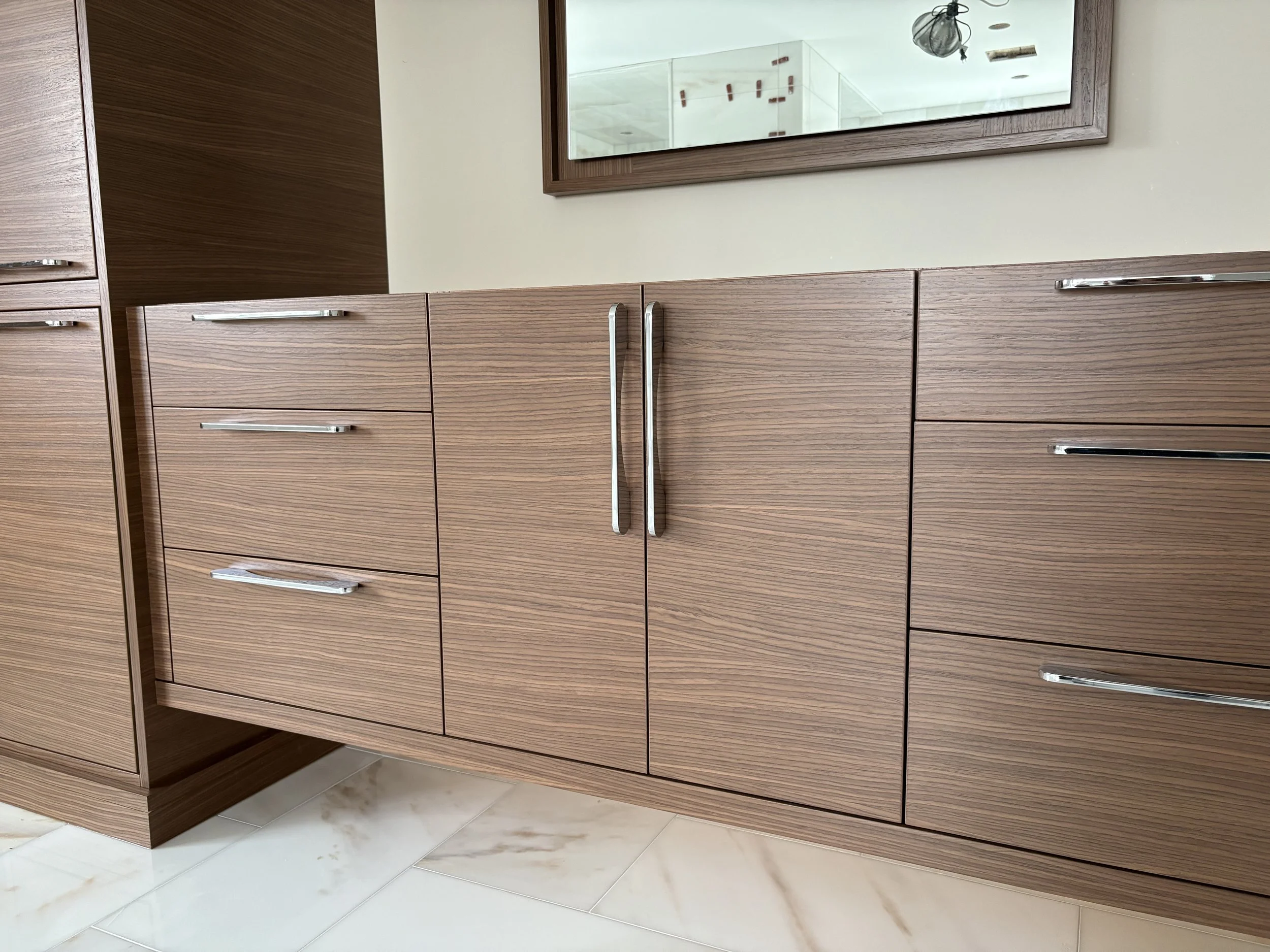 A room with wooden cabinets and drawers, two large mirrors on the wall, one window reflecting sunlight, and a tiled floor.