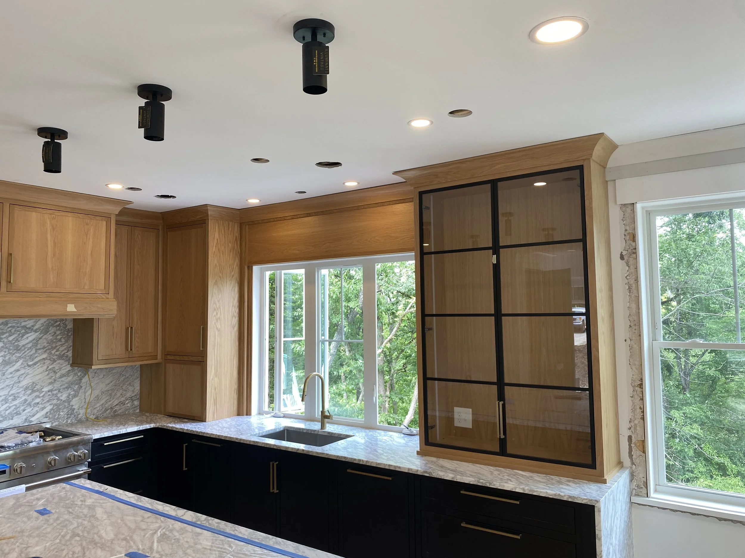 Kitchen with wooden upper cabinets, black lower cabinets, marble countertops, a window above the sink, and a black-framed glass cabinet. The ceiling has recessed lighting and unfinished wiring for additional lighting.