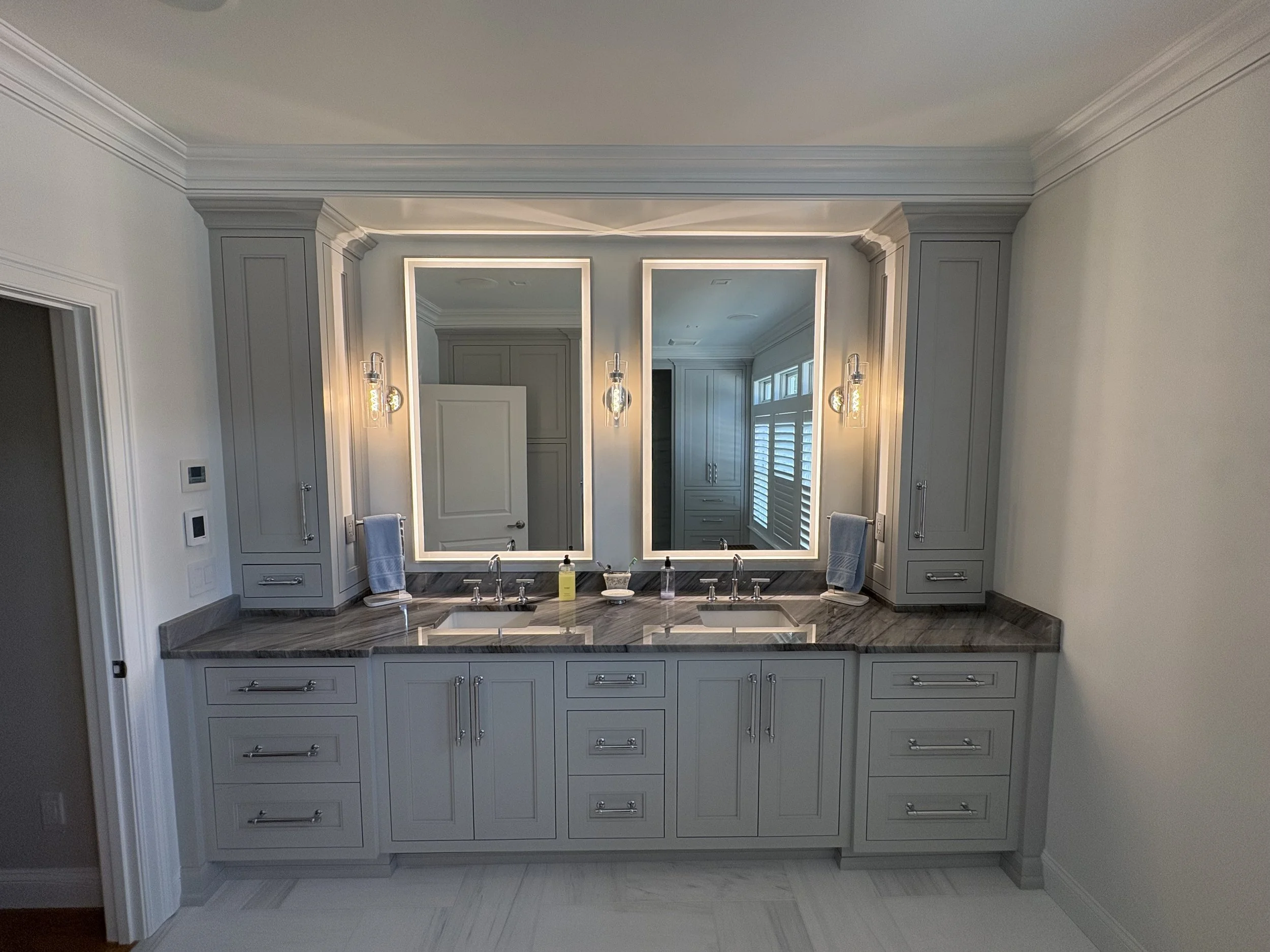 A modern bathroom vanity with two illuminated mirrors, two sinks, gray cabinetry, and a marble countertop.