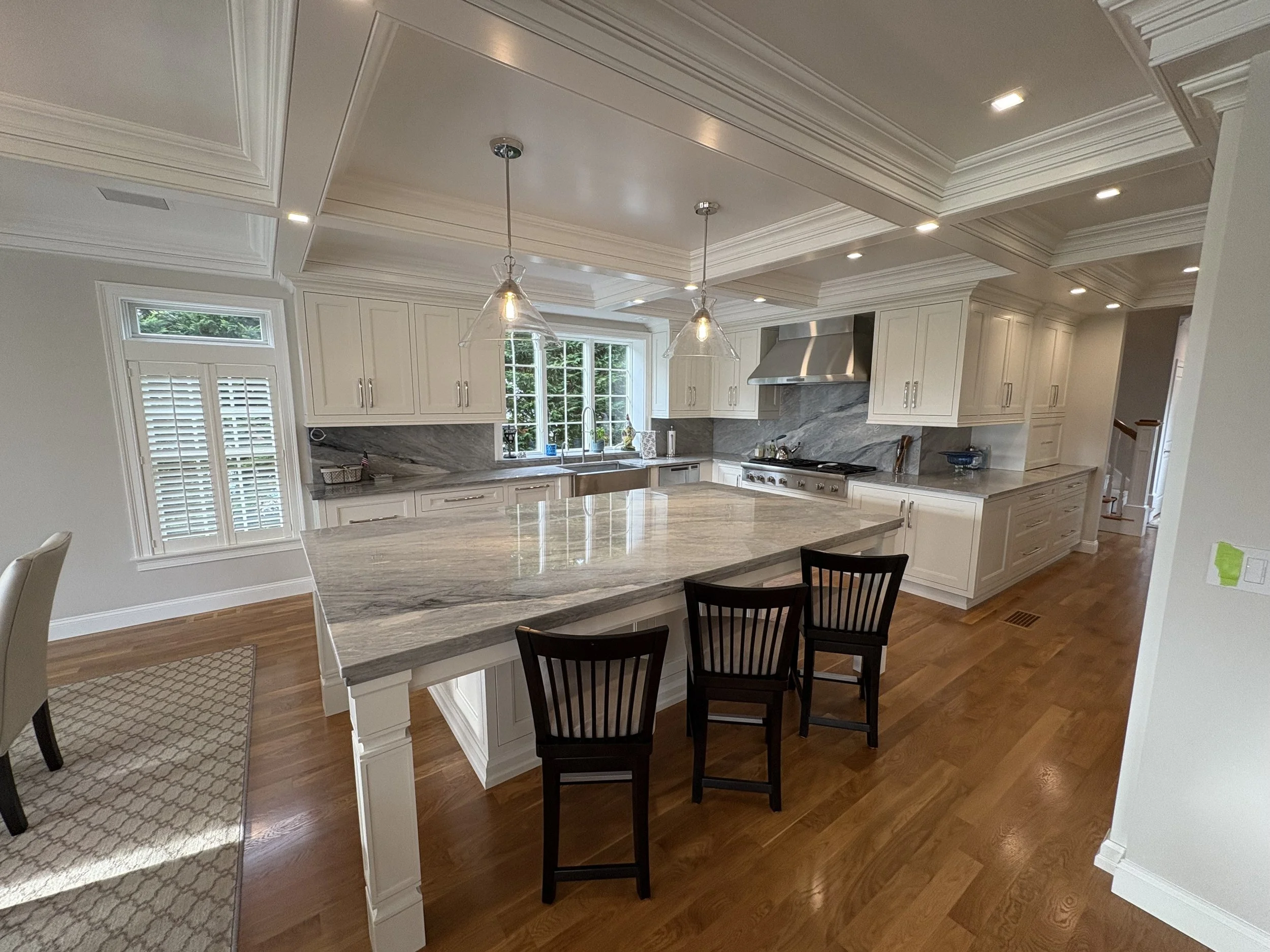 Spacious kitchen with white cabinets, gray marble countertops, and a large central island. There are two pendant lights above the island, a window above the sink, and hardwood floors.