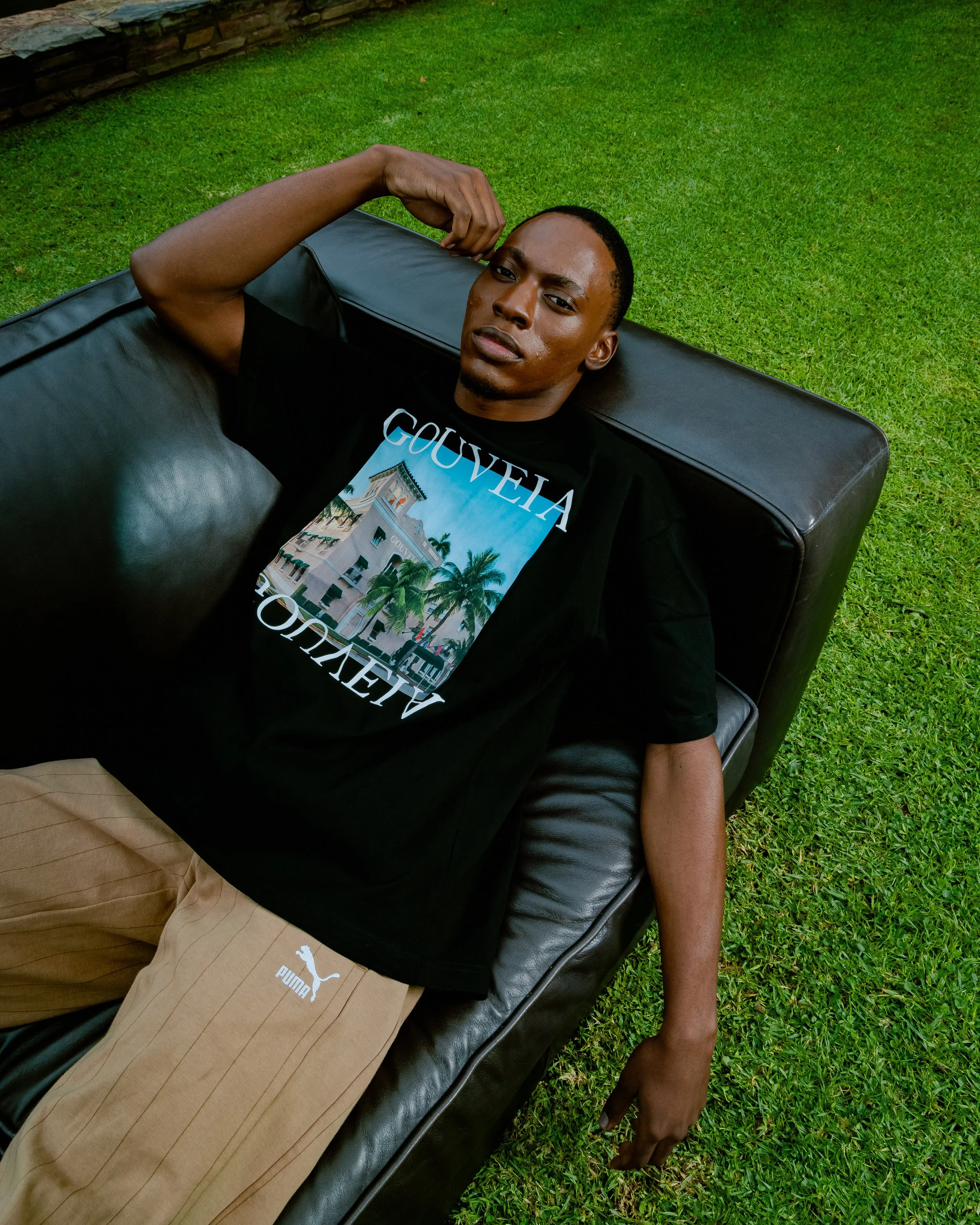 A young man laying on a black leather couch outdoors on green grass, wearing a black T-shirt with a graphic of a building and trees, tan shorts, and resting his head on the couch, with one arm bent and hand near his temple, looking at the camera with a relaxed expression.