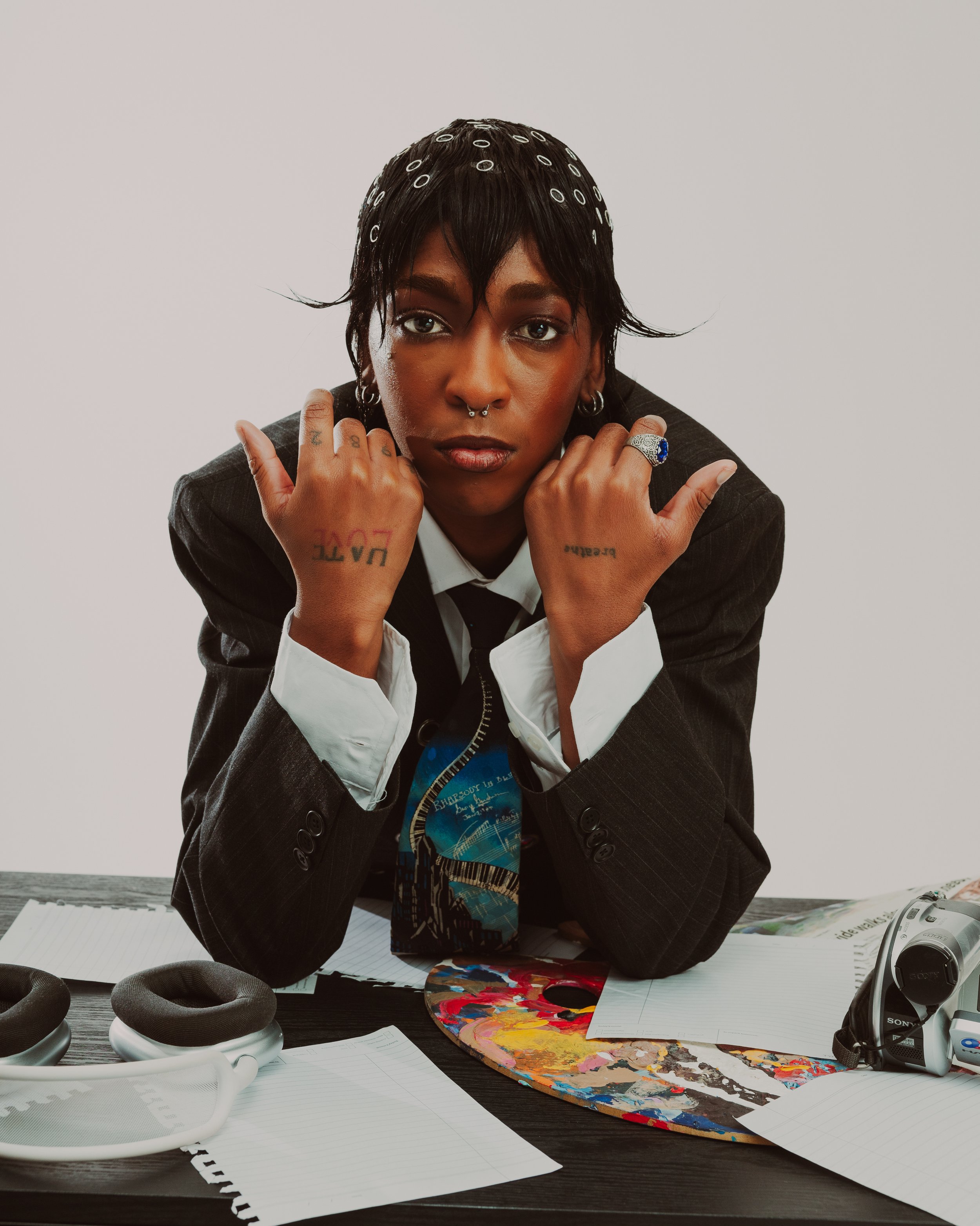 A person with dark hair and tattoos on their hands sits at a table surrounded by headphones, papers, a paint palette, and art supplies. They have a septum piercing and are wearing a dark blazer and a white shirt. The background is plain and light-colored.