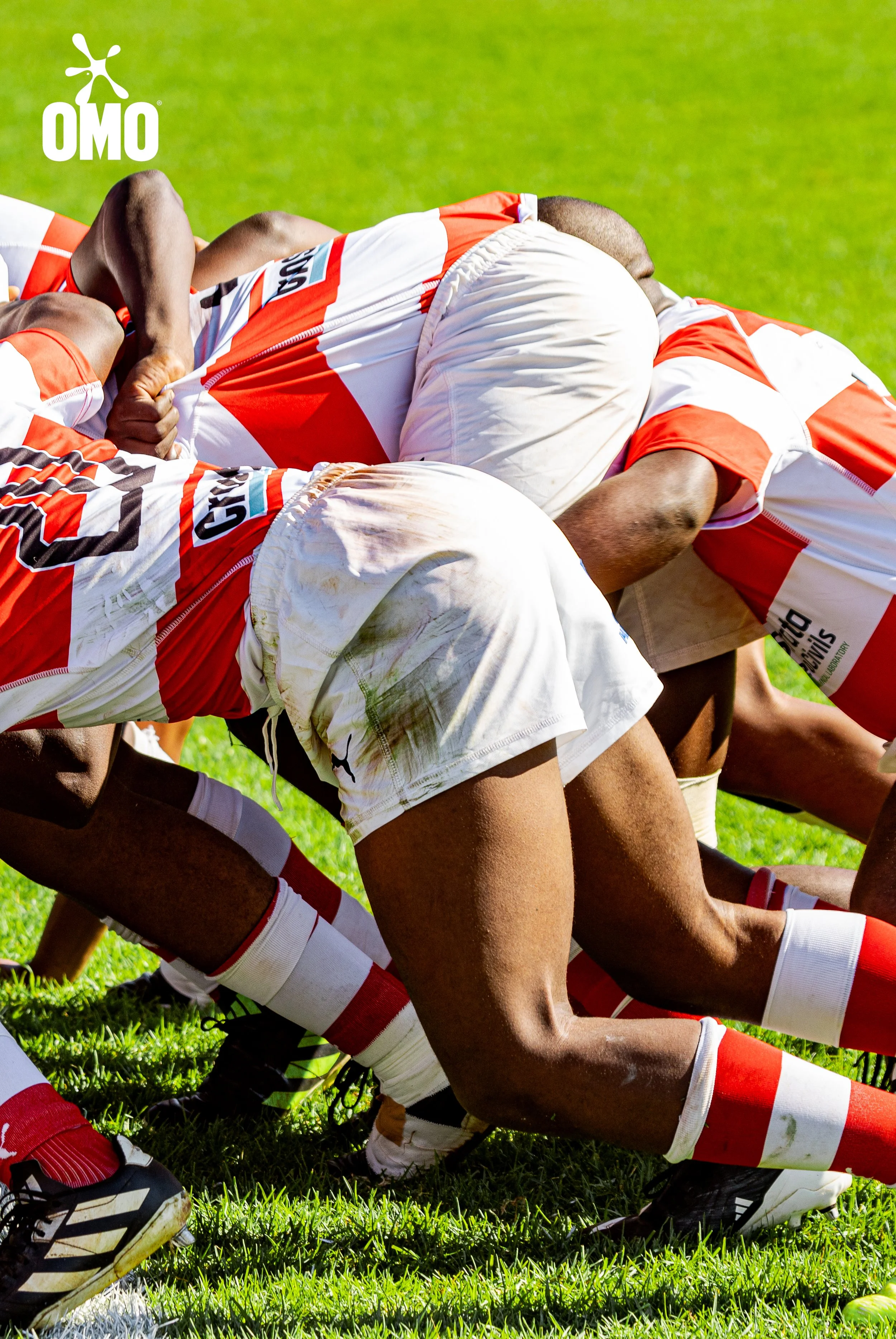 Rugby players in a scrum on a grassy field, wearing red and white uniforms with socks and cleats.