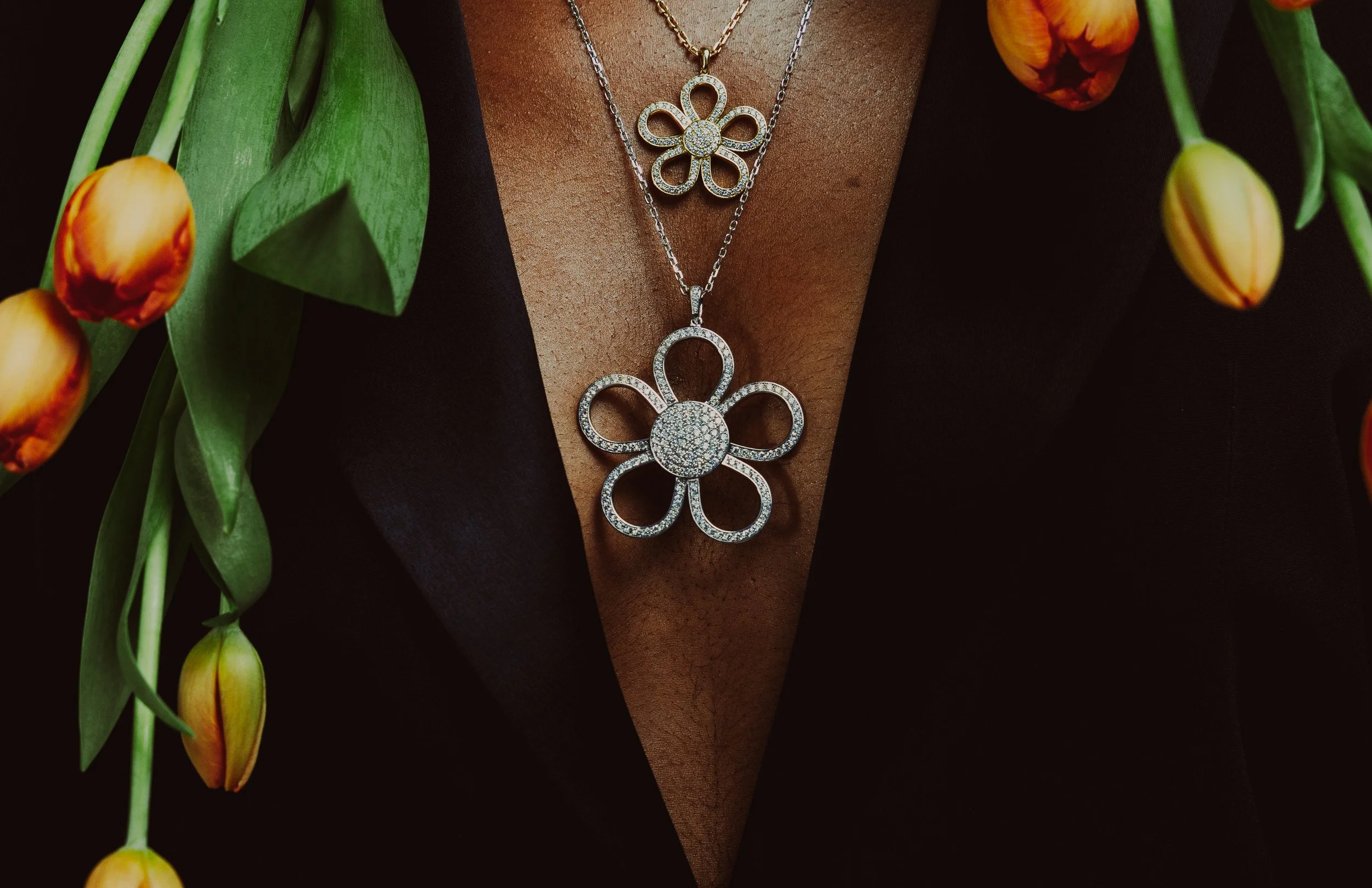 Close-up of a person wearing layered silver necklaces with large flower pendants encrusted with diamonds, surrounded by orange tulips and green leaves.