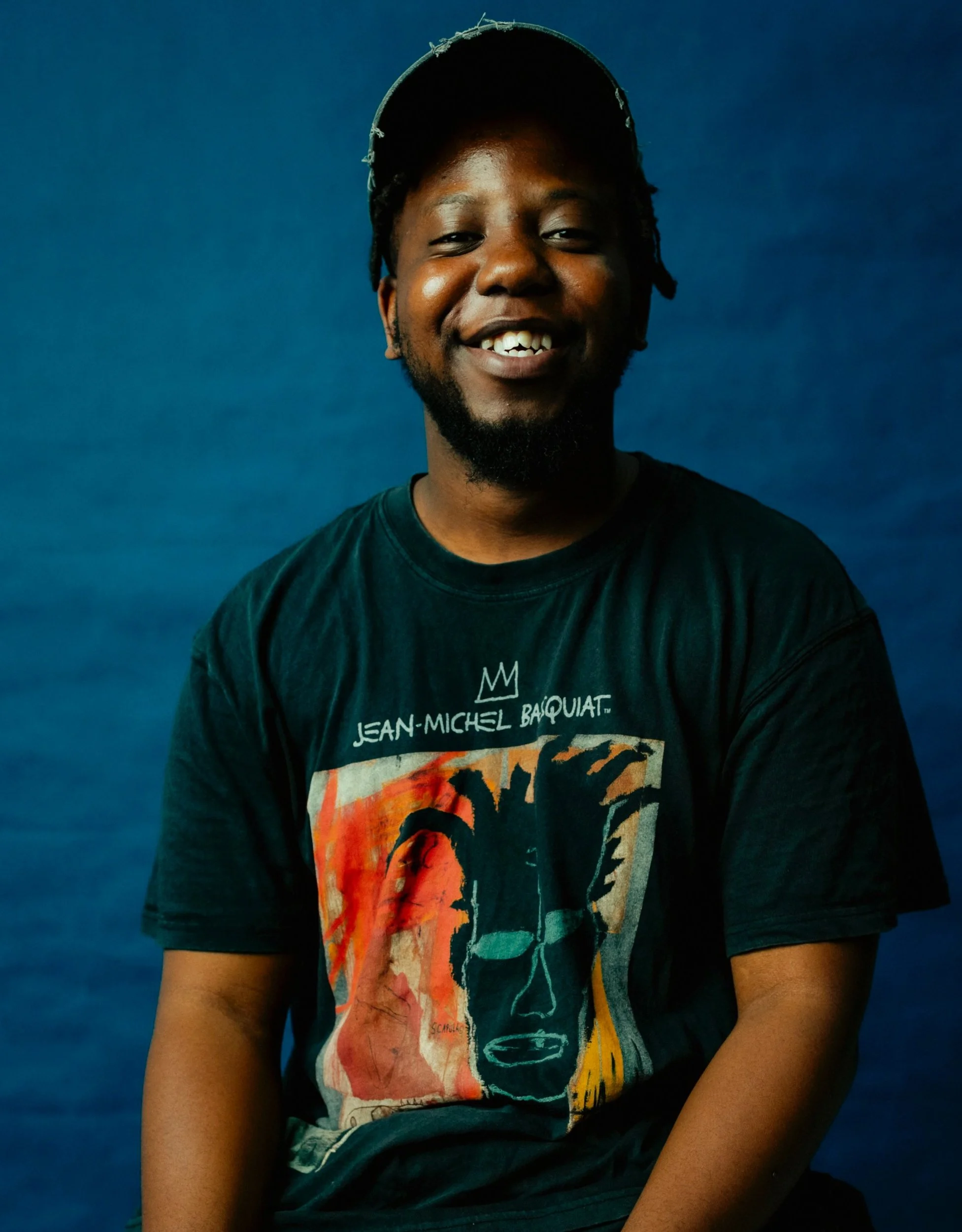 A young man with dark skin, wearing a black Jean-Michel Basquiat t-shirt and a cap, is smiling against a blue background.