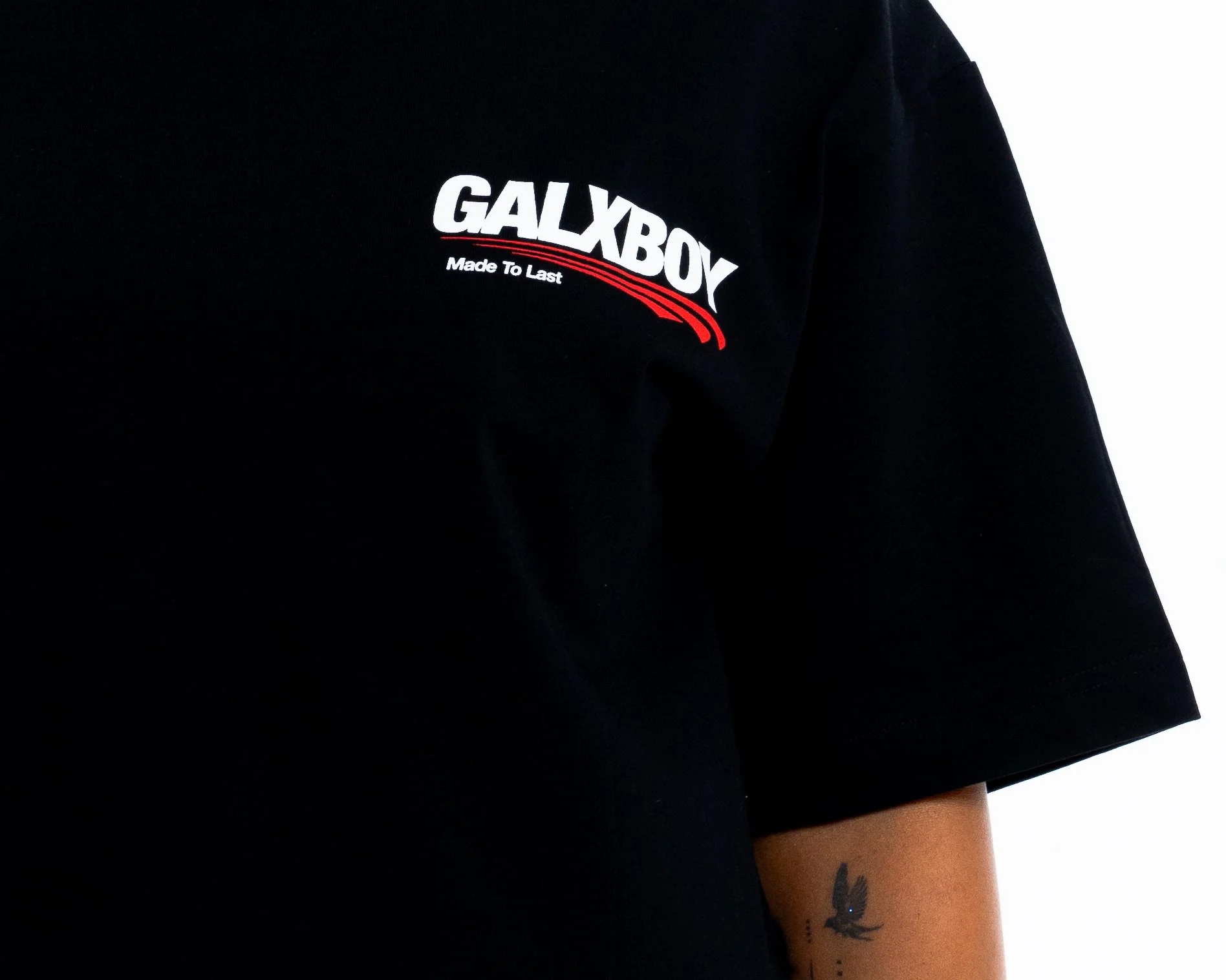 Close-up of person wearing a black shirt with a logo that reads "GALAXBOX" and "Made To Last" underneath. Part of person's arm shows a tattoo of a bird in flight.
