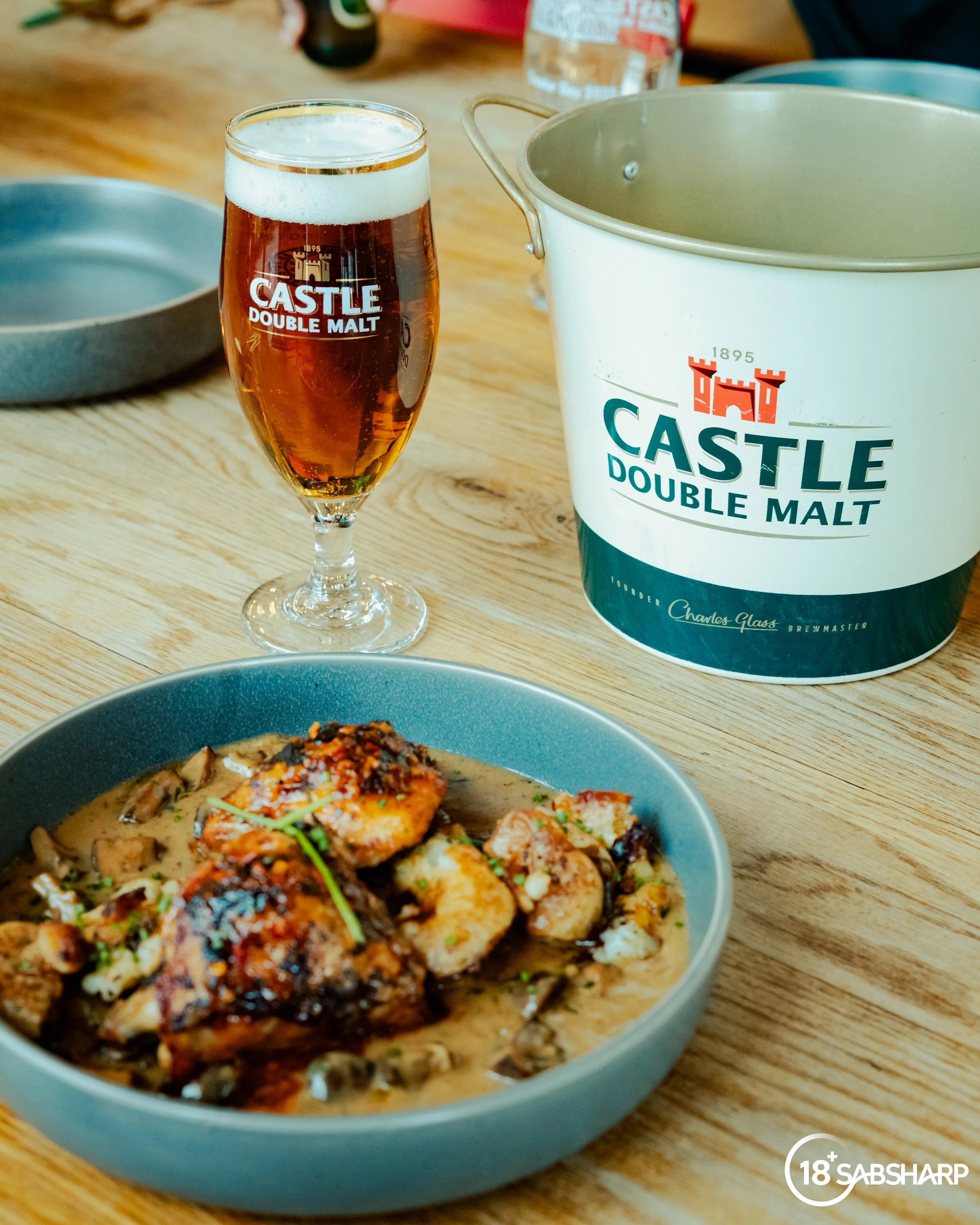 A glass of amber beer labeled 'Castle Double Malt' next to a bucket with the same label, and a bowl of seafood with sauce on a wooden table.