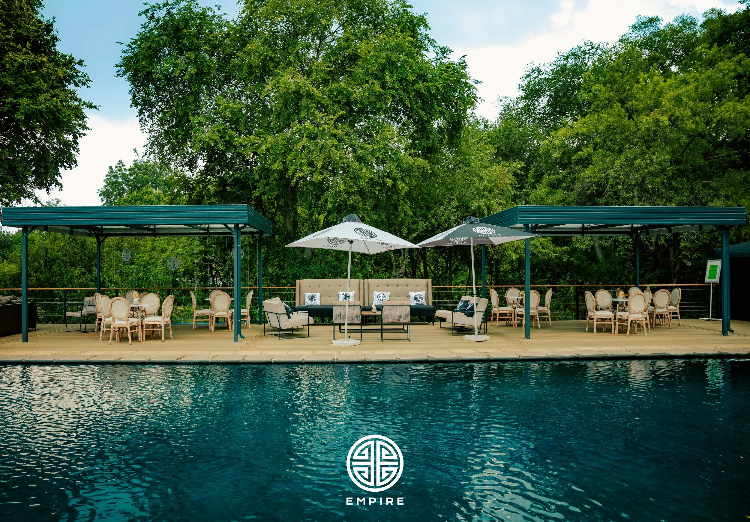 Luxury outdoor poolside seating area with tables, chairs, umbrellas, and a seating lounge on a wooden deck surrounded by green trees