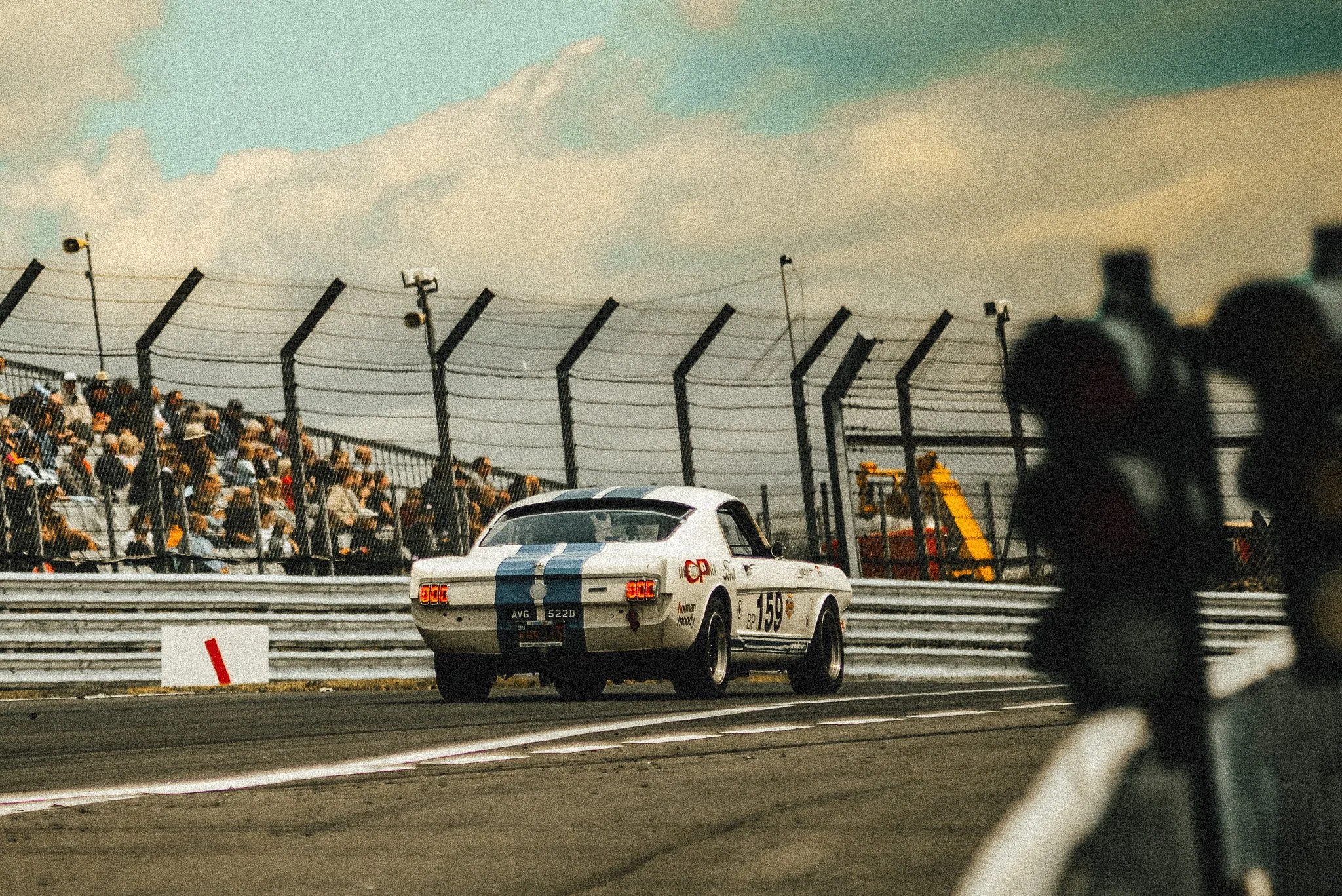 Vintage race car on a racetrack with a crowd watching from bleachers, fenced race track, and sky in the background.