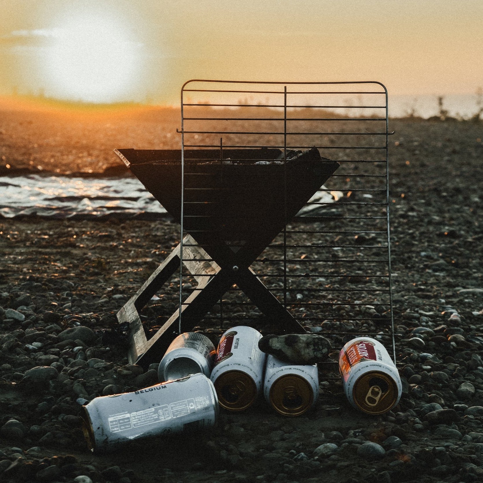 A small charcoal barbecue grill on a pebble beach at sunset, with several empty beer cans lying on the ground nearby.