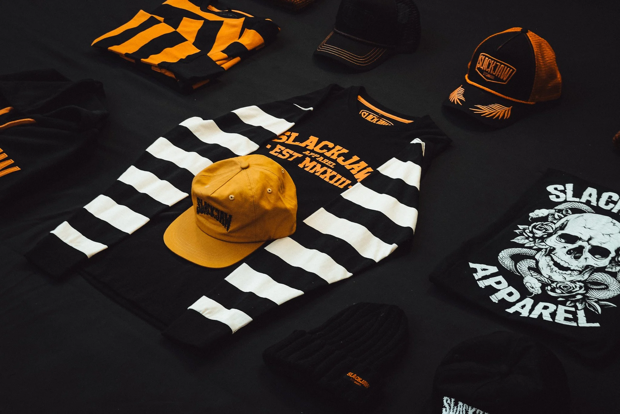 Display of black and white striped long-sleeve shirt, titled 'SLACKJAW,' and accessories including hats, a T-shirt with skull and snake design, and beanie, all featuring the 'SLACKJAW' branding in black and orange on a black surface.