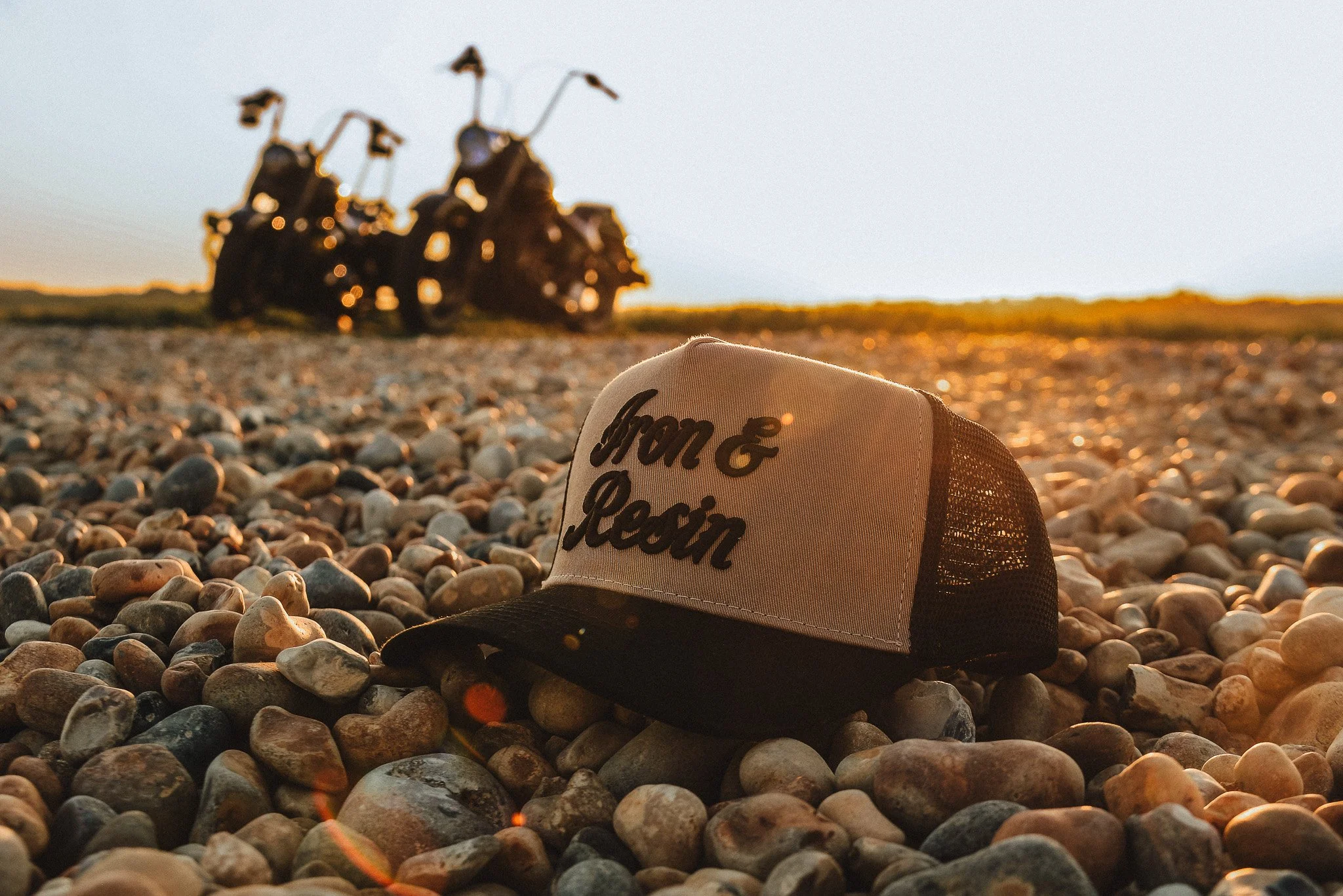 A black and white trucker hat with the words 'Born & Reconstructed' rests on a rocky beach, with several motorcycles blurred in the background during sunset.