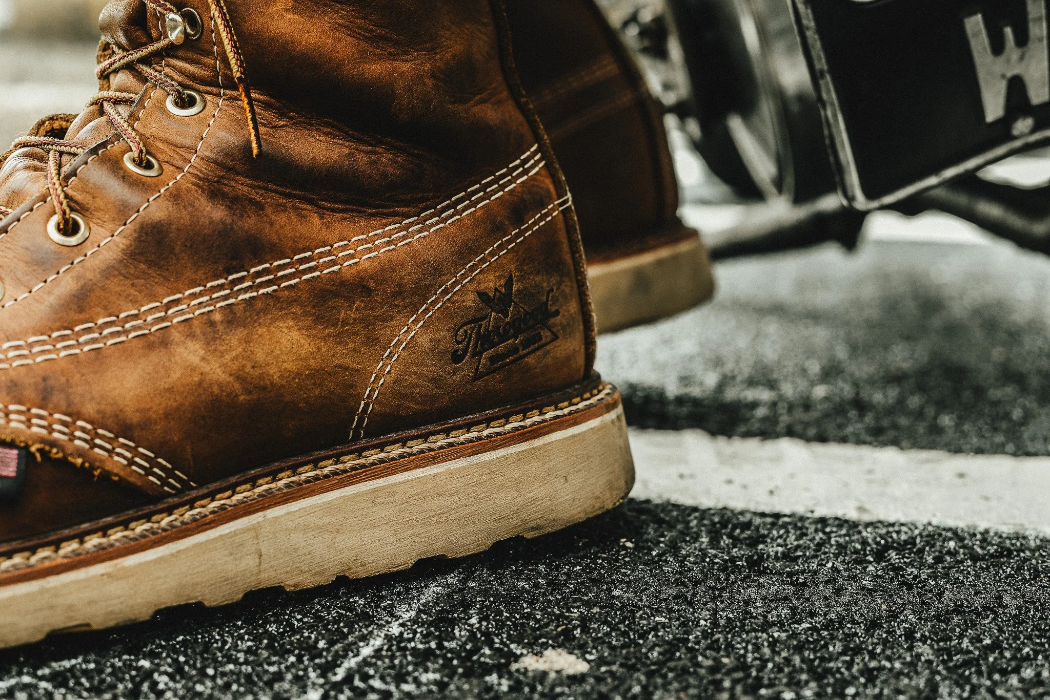Close-up of a brown leather Timberland boot on rough asphalt ground, showing detailed stitching and the Timberland logo.