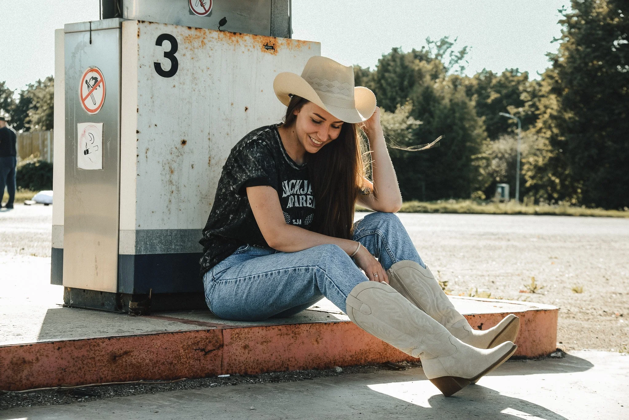 A young woman wearing cowboy boots, a wide-brimmed hat, a black graphic t-shirt, and blue jeans, sitting on the sidewalk next to a rusted electrical box outdoors during daytime, smiling and looking down.