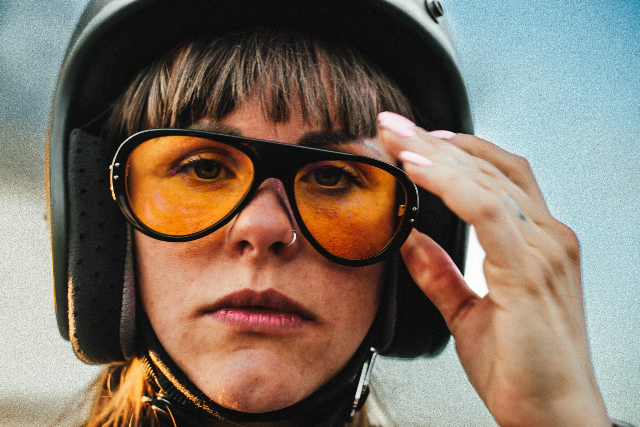 A woman wearing a helmet and orange-tinted sunglasses, adjusting her glasses with her hand.