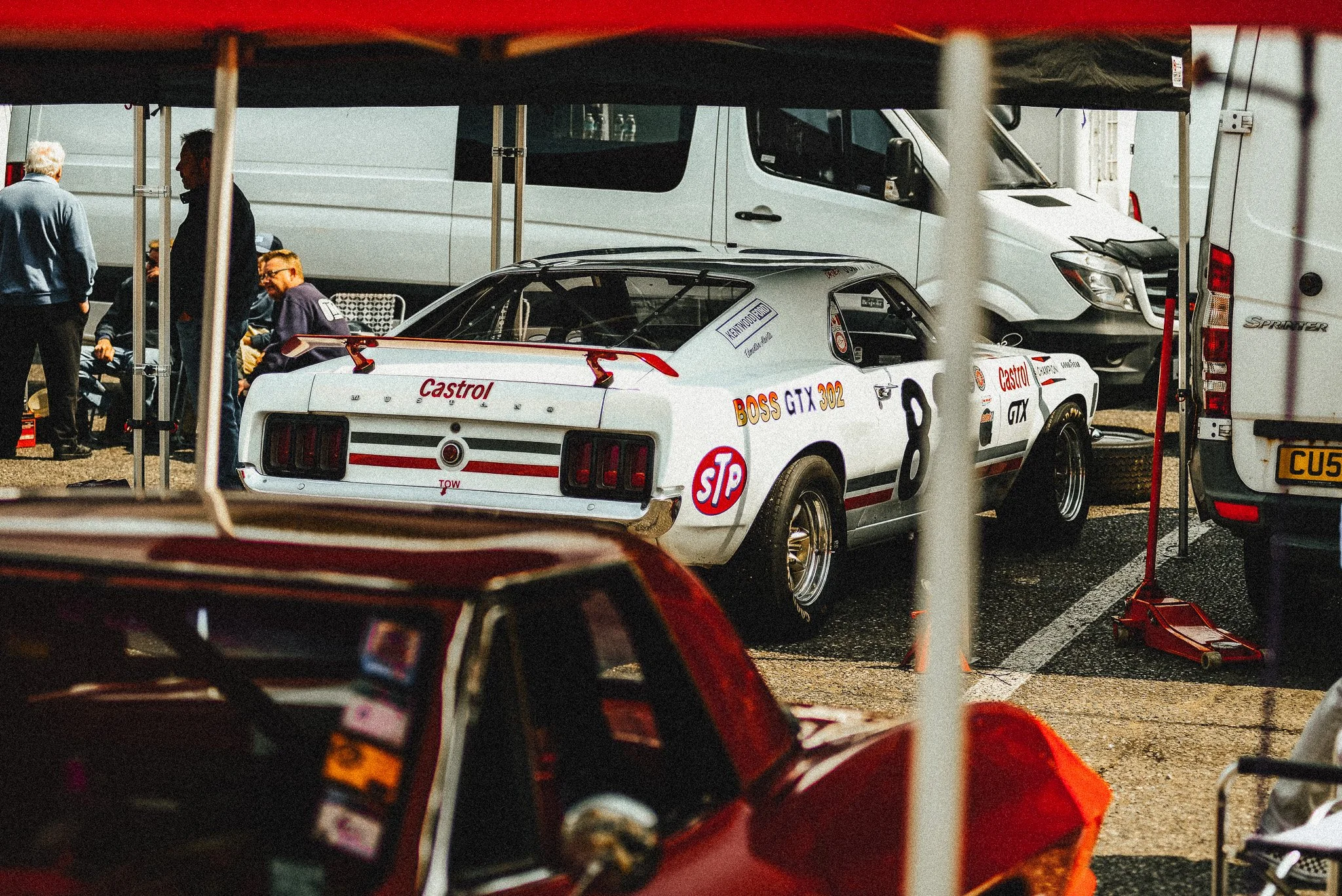 A vintage racing car with racing decals and the number 8 parked in a paddock, seen through a partially obscured view with tents and other vehicles around.