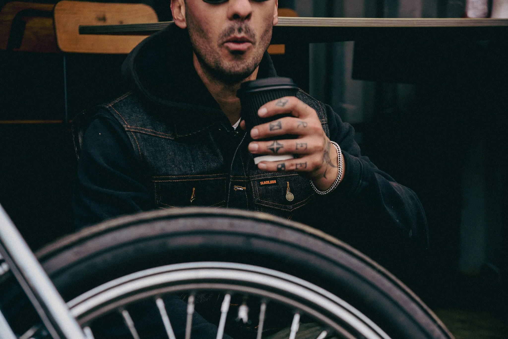 A man with tattoos on his fingers and hand holding a black coffee cup, wearing a denim jacket and dark sunglasses.