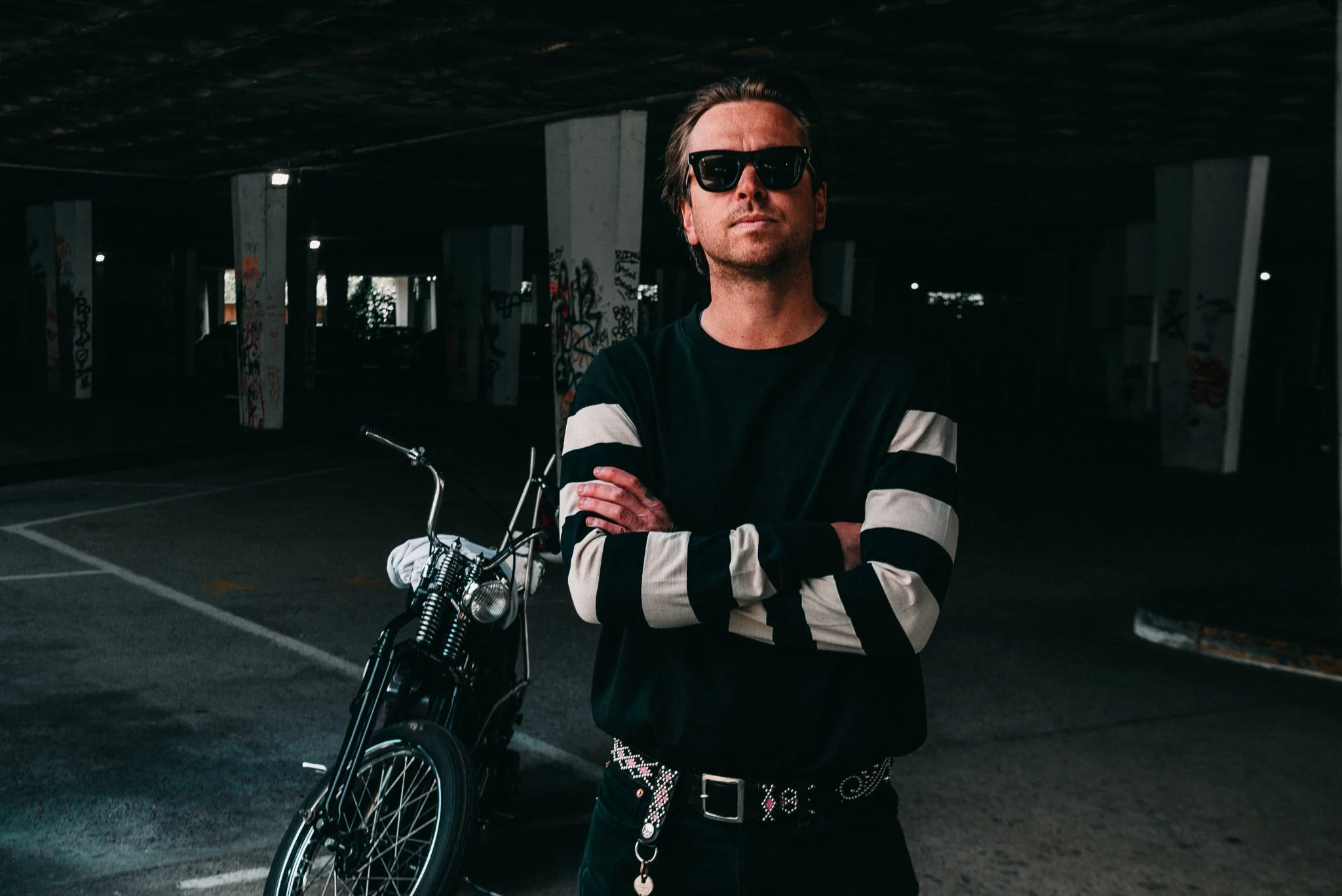 A man with dark sunglasses, wearing a black and white striped long-sleeve shirt, standing arms crossed next to a motorcycle in an underground parking garage with graffiti on the pillars.